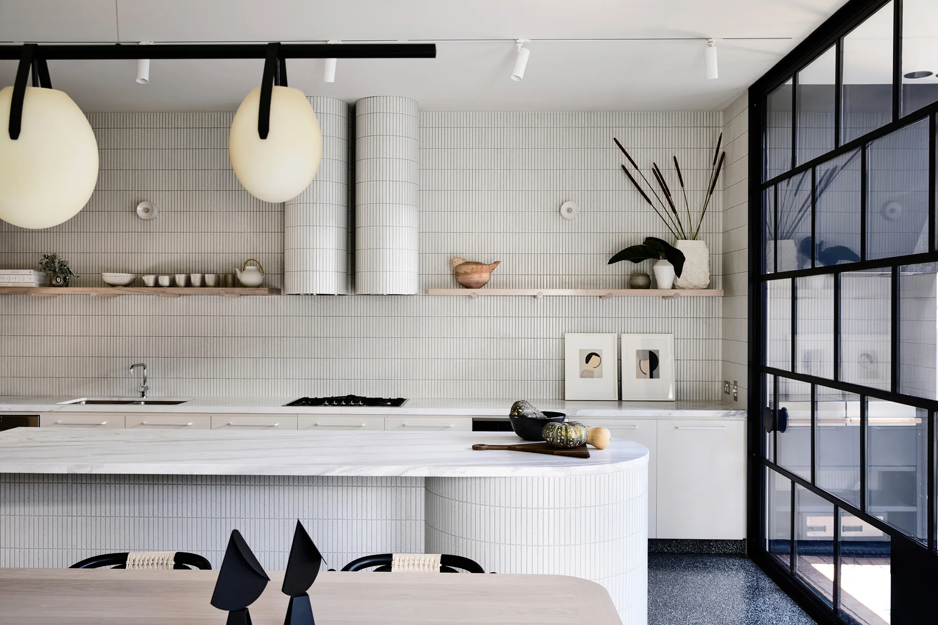 A modern kitchen featuring a curved white marble island and vertical-tiled backsplash walls with integrated cylindrical range hoods.