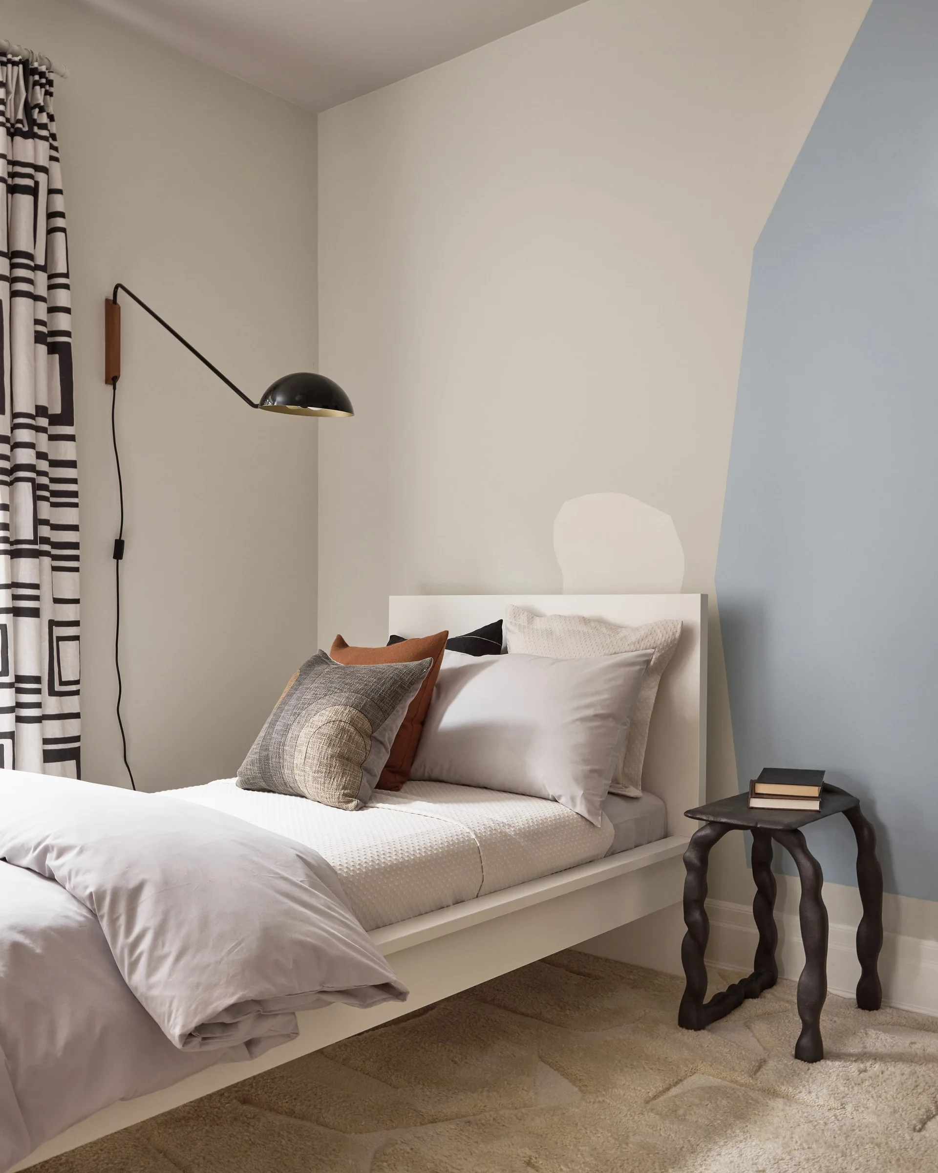 A modern bedroom featuring a white bed with layered pillows, a black swing-arm wall sconce, and a blue painted geometric accent wall.