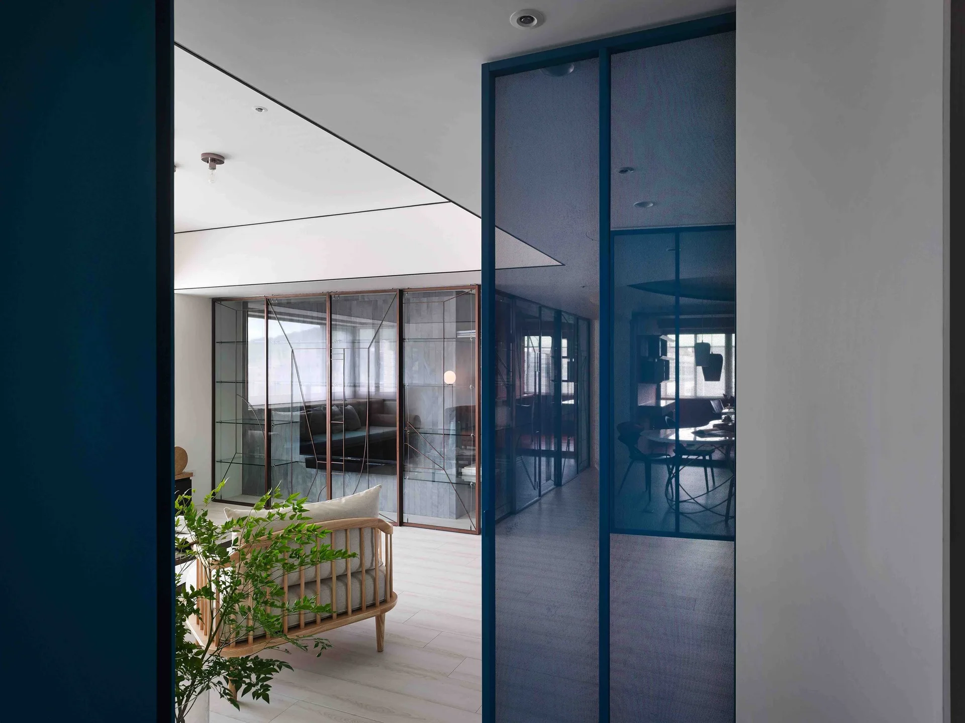 A modern apartment living area featuring a blue-framed sliding glass partition, light wood spindle sofa, and bronze metal-framed glass dividers.