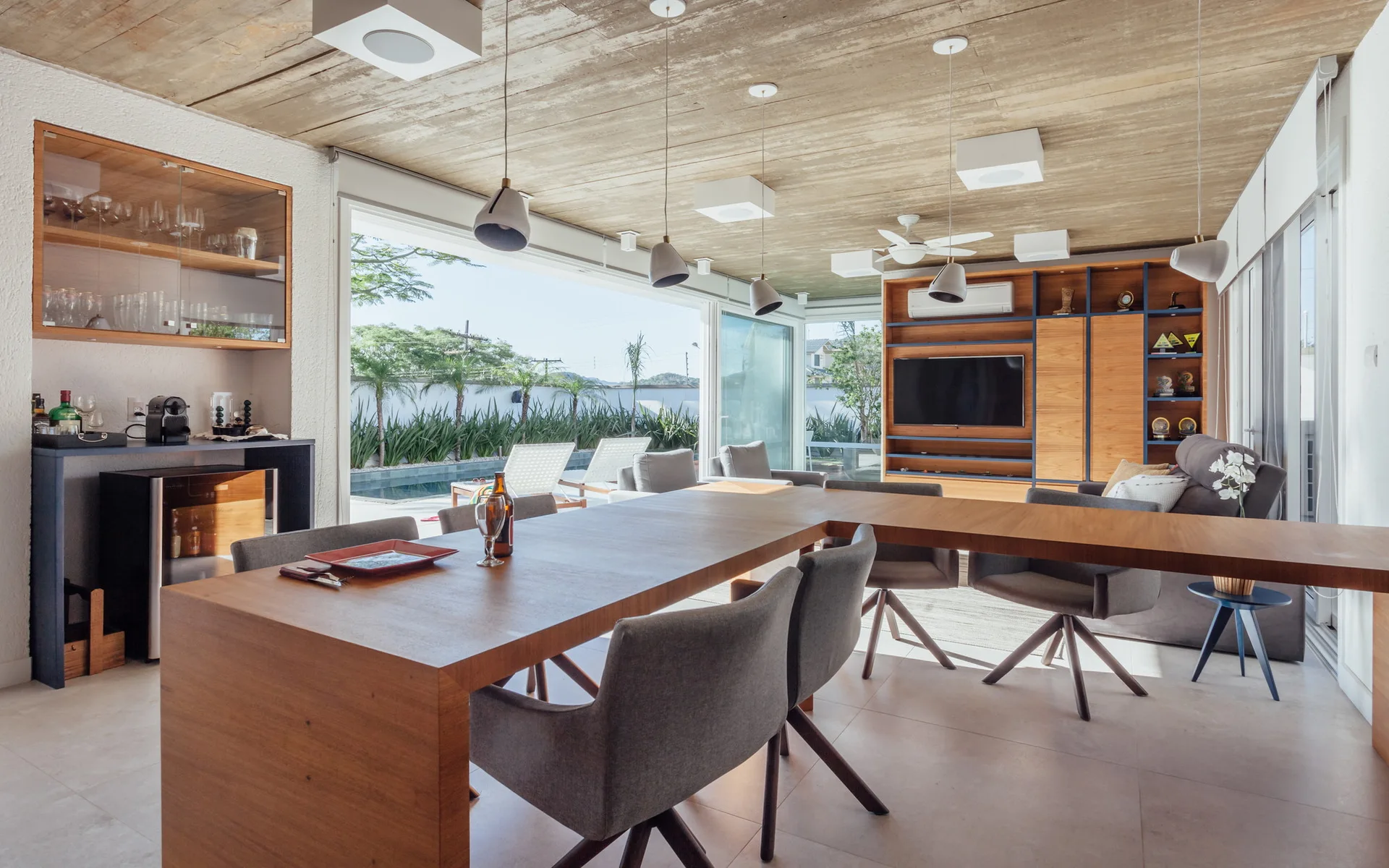 An open-plan dining and living area features a long wooden table, grey chairs, a concrete ceiling, and glass sliding doors opening to an outdoor pool.