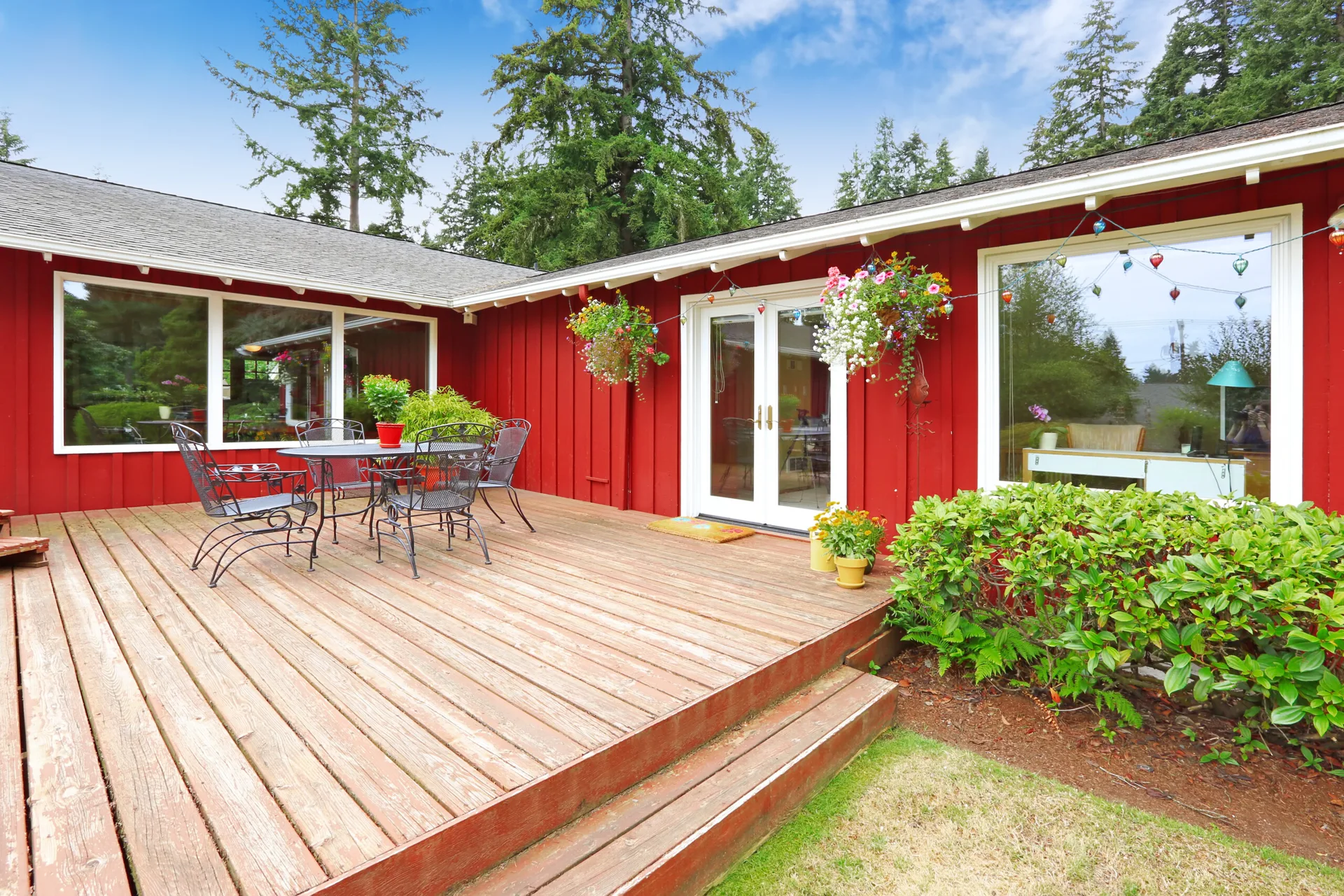 A spacious wooden deck featuring a black metal patio table and chairs set against vibrant red vertical siding with white-trimmed windows.