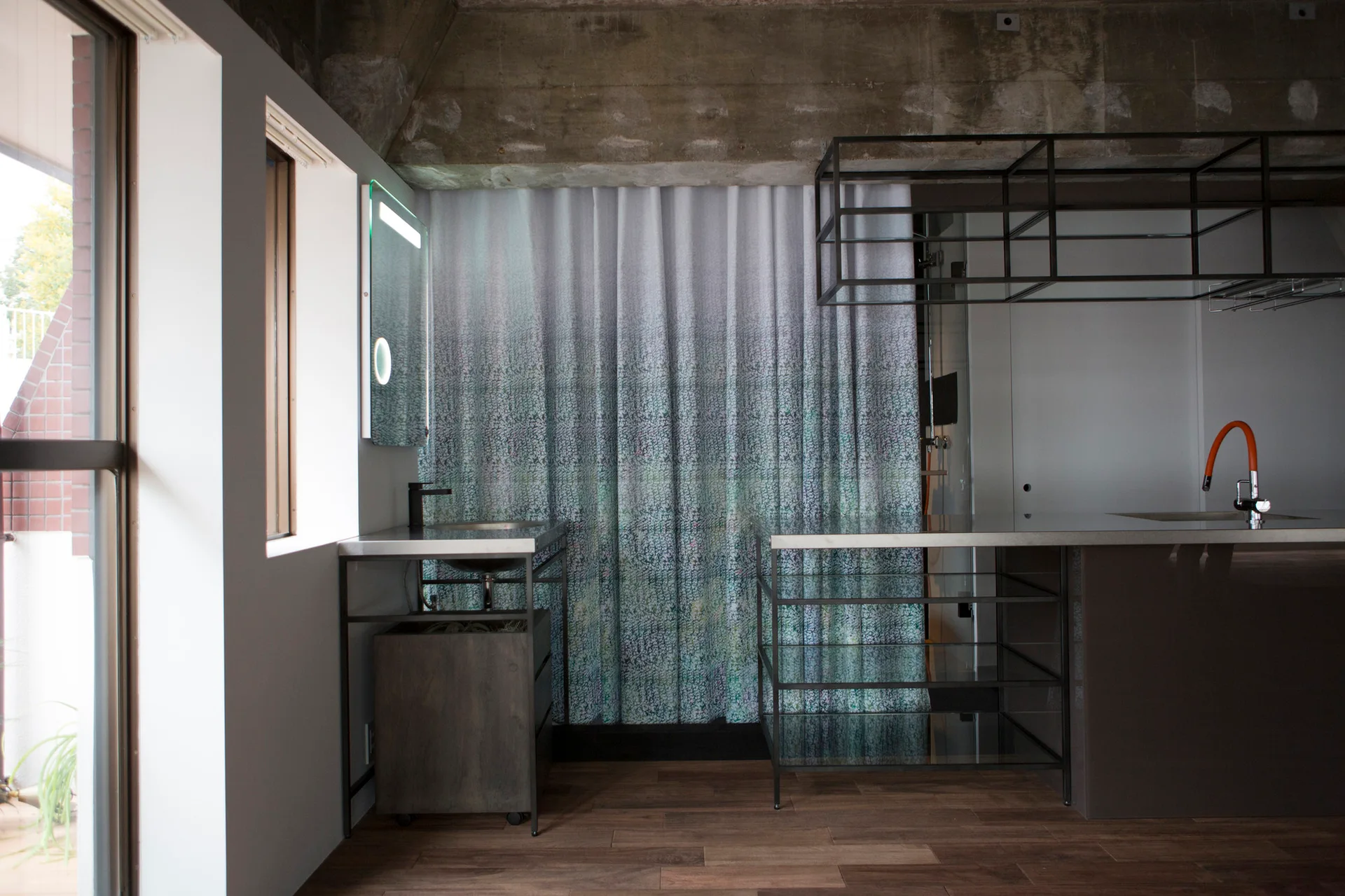 An industrial bathroom features an exposed concrete ceiling, a metal-framed vanity with an LED mirror, and a green ombre curtain.