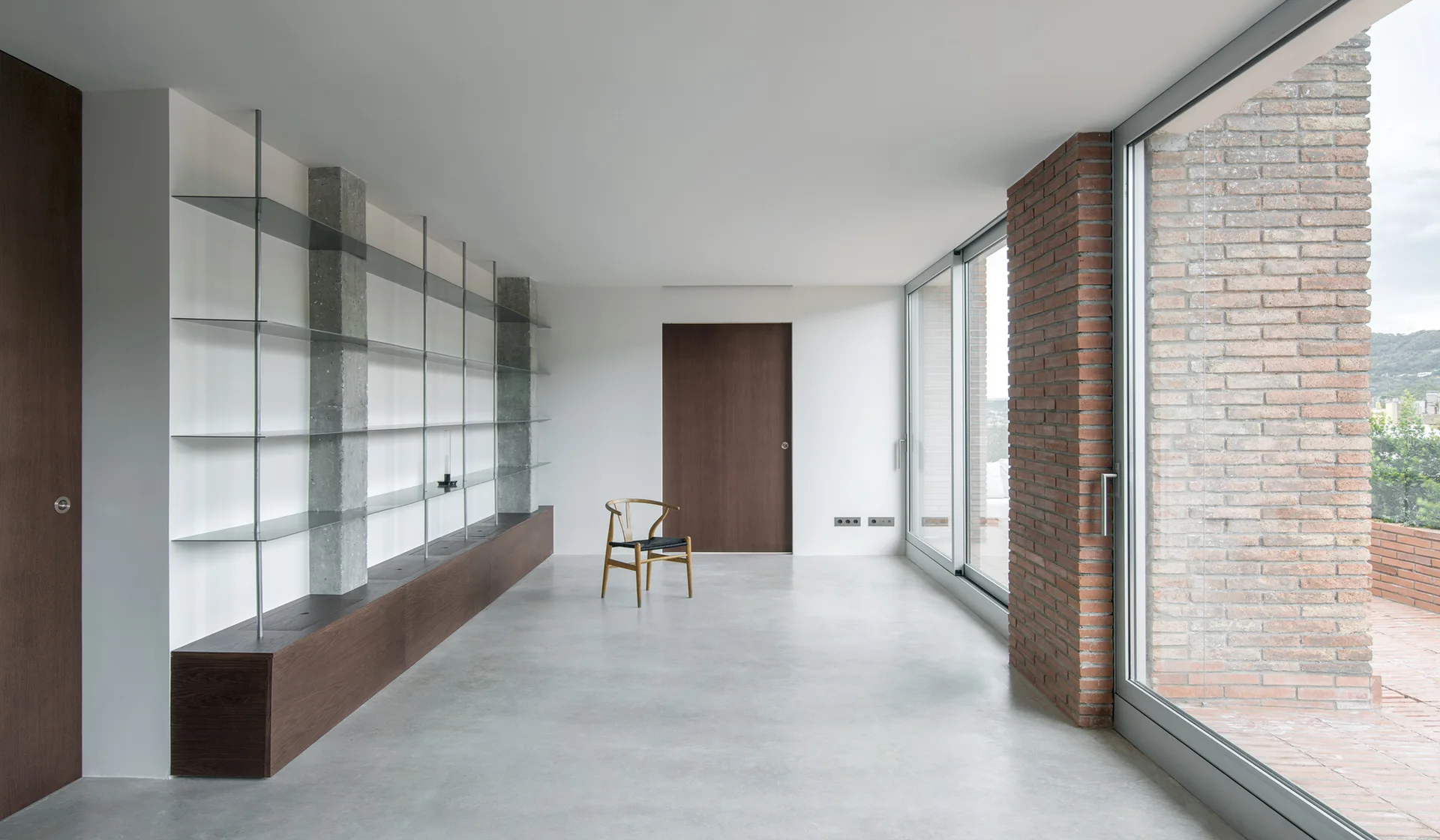A minimalist room features a poured concrete floor, a low wood cabinet with glass shelves, a single wooden chair, and large glass patio doors.