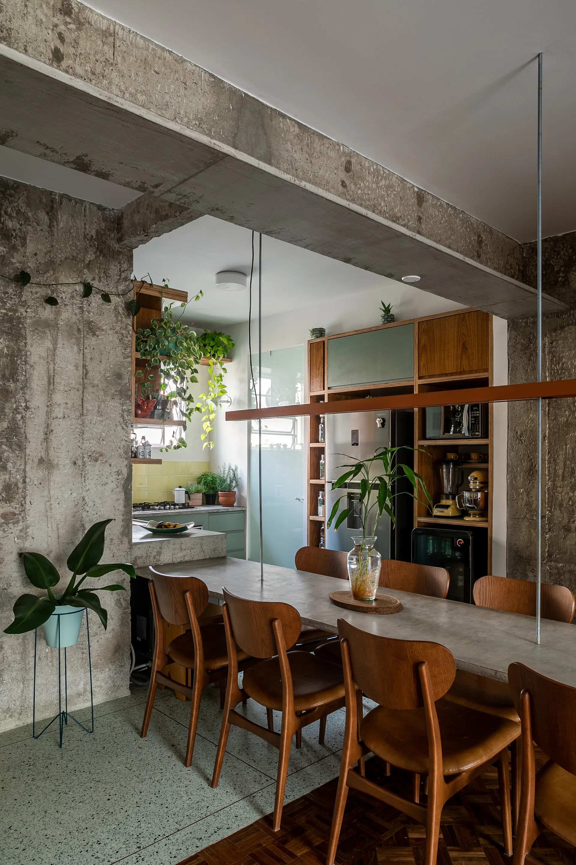 A dining table with wooden chairs stands beneath an exposed concrete beam in an open room next to a kitchen with wooden cabinets.