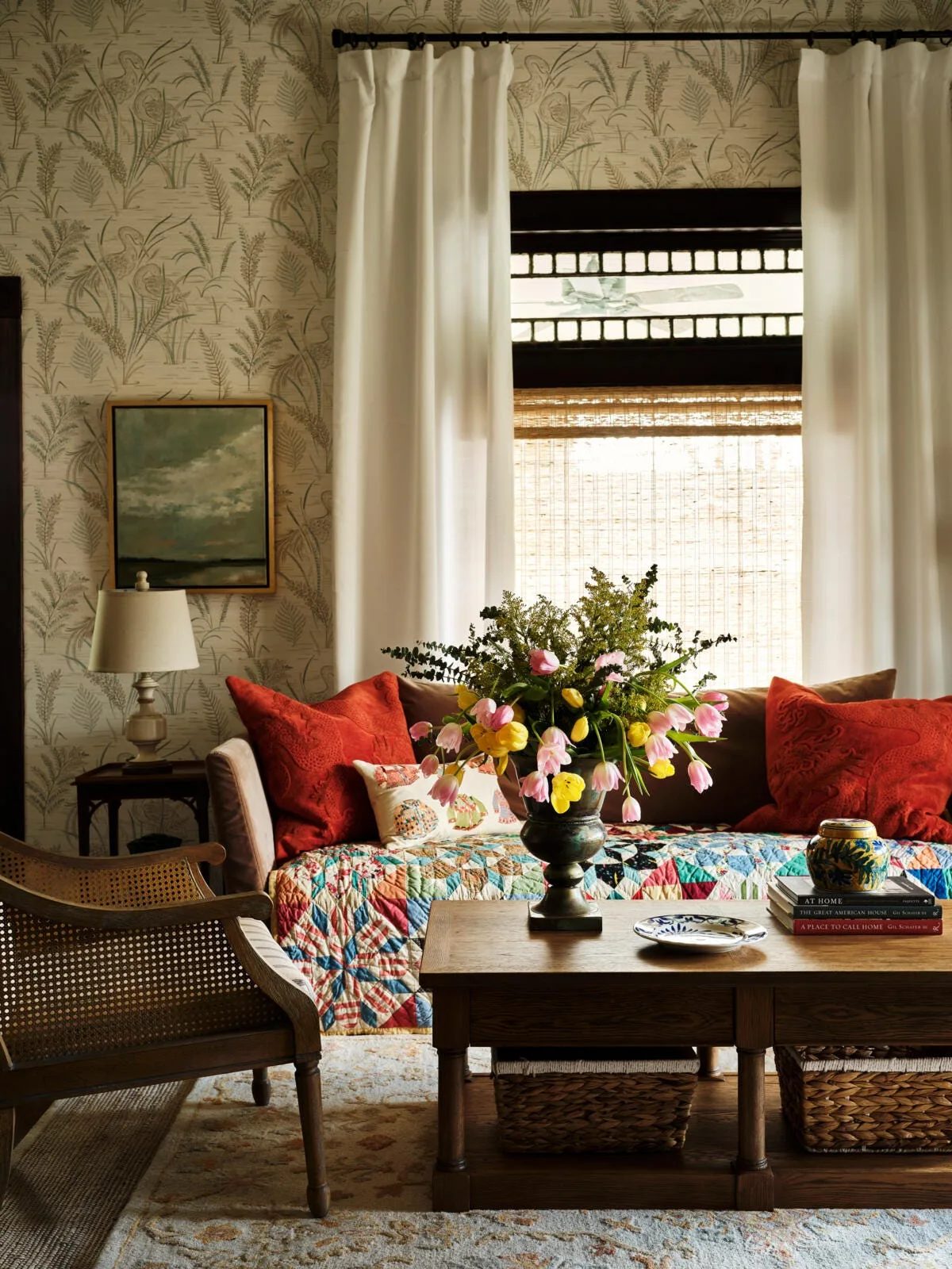 A traditional living room featuring botanical fern wallpaper, a wood coffee table with tulips, and a sofa with a colorful patchwork quilt.