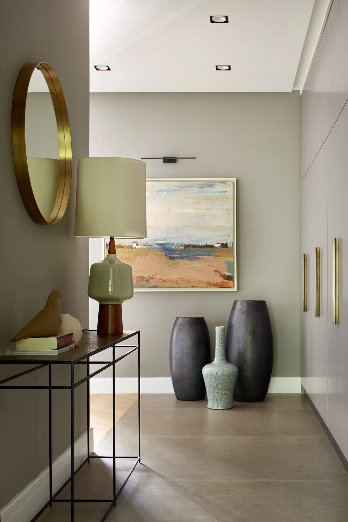 A modern grey hallway features a black metal console table with a ceramic lamp, large dark floor vases, and built-in minimalist storage cabinets.