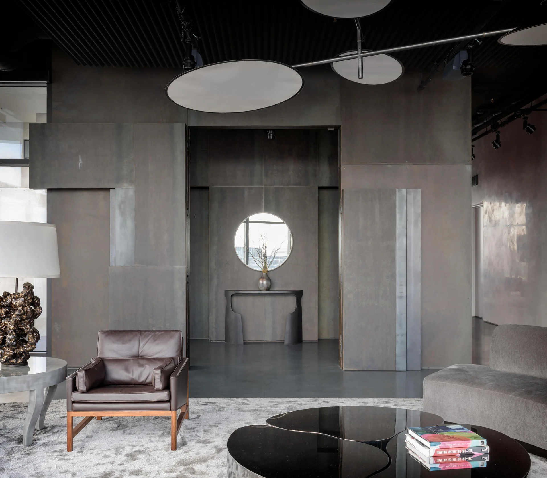 A modern living area features dark charcoal metal wall panels, a black slatted ceiling, a brown leather armchair, and an organic-shaped black coffee table.