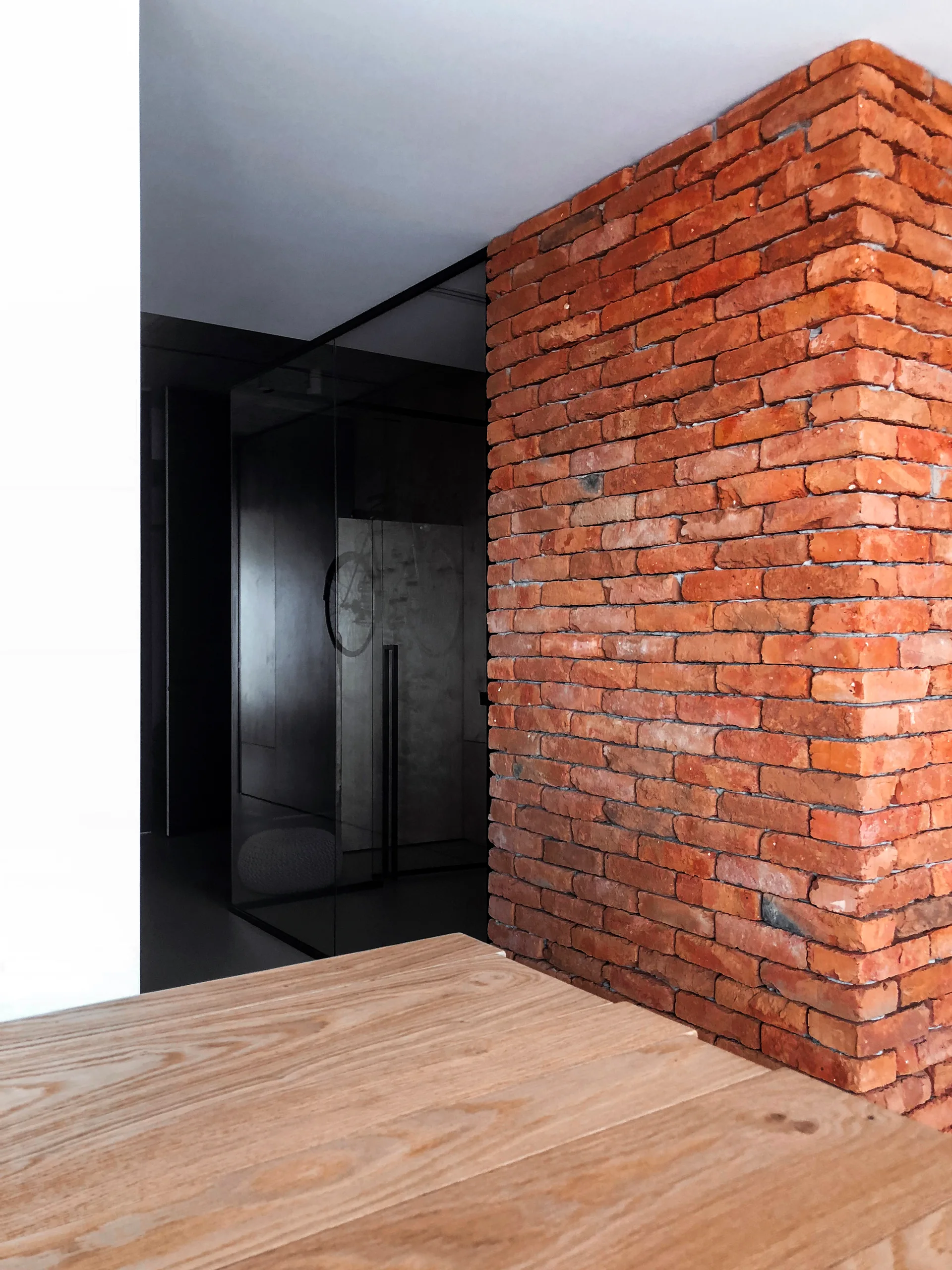 A modern minimalist interior featuring an exposed red brick corner wall next to a dark glass partition and a light wood table.