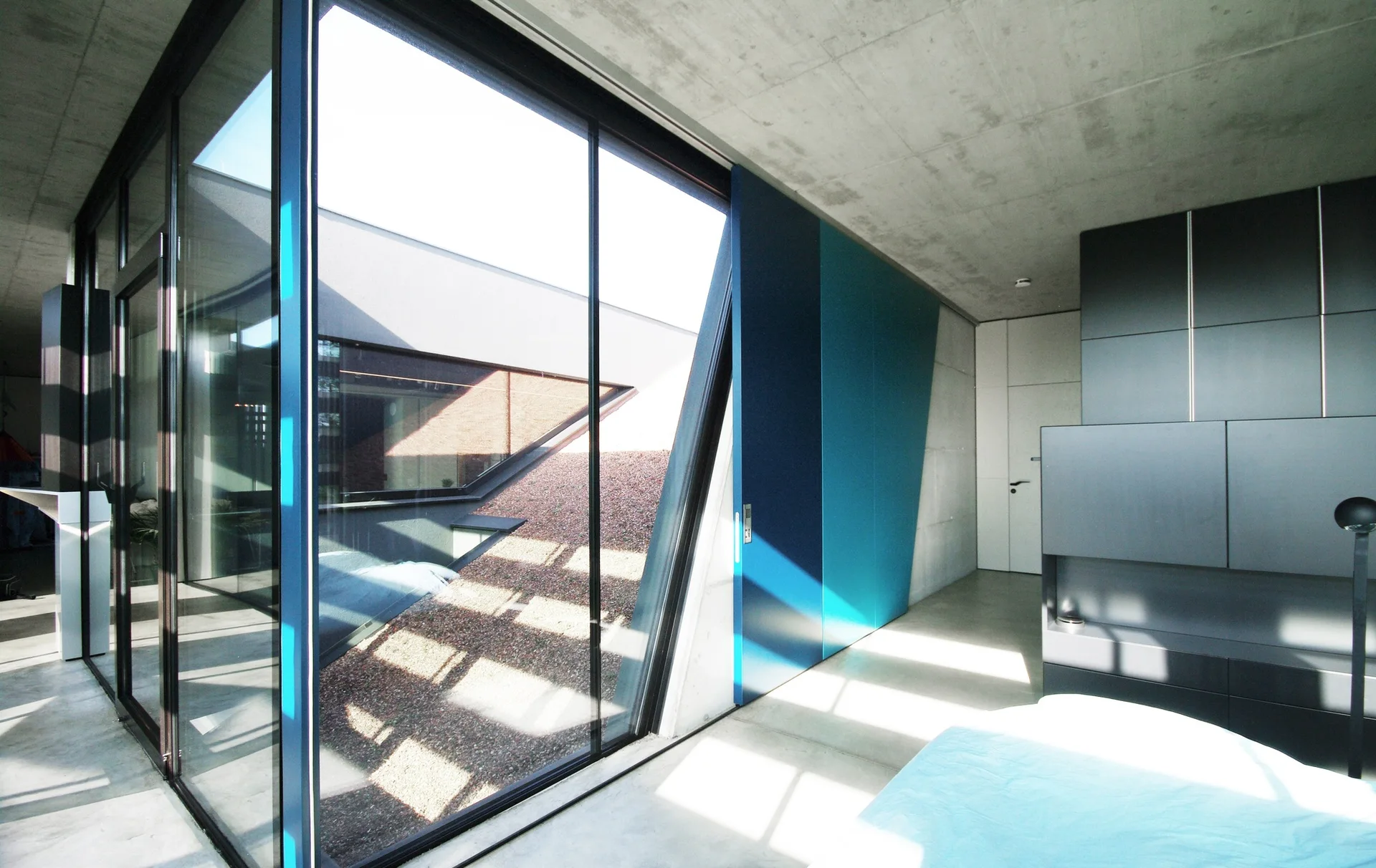 A minimalist room with exposed concrete walls and ceiling, a large glass partition overlooking a gravel courtyard, and a blue sliding door.