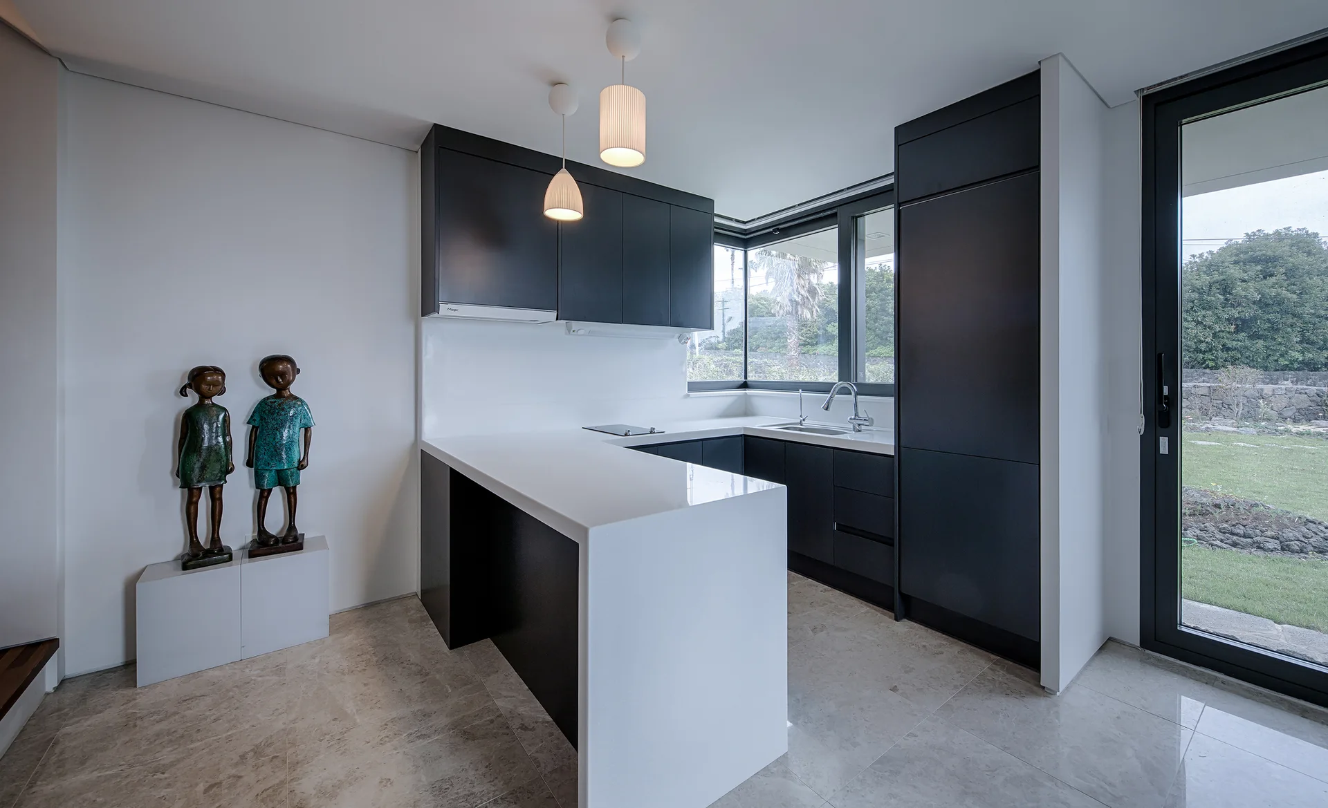A minimalist kitchen features matte black cabinets, a white waterfall countertop peninsula, a corner window, and two small bronze-like statues.