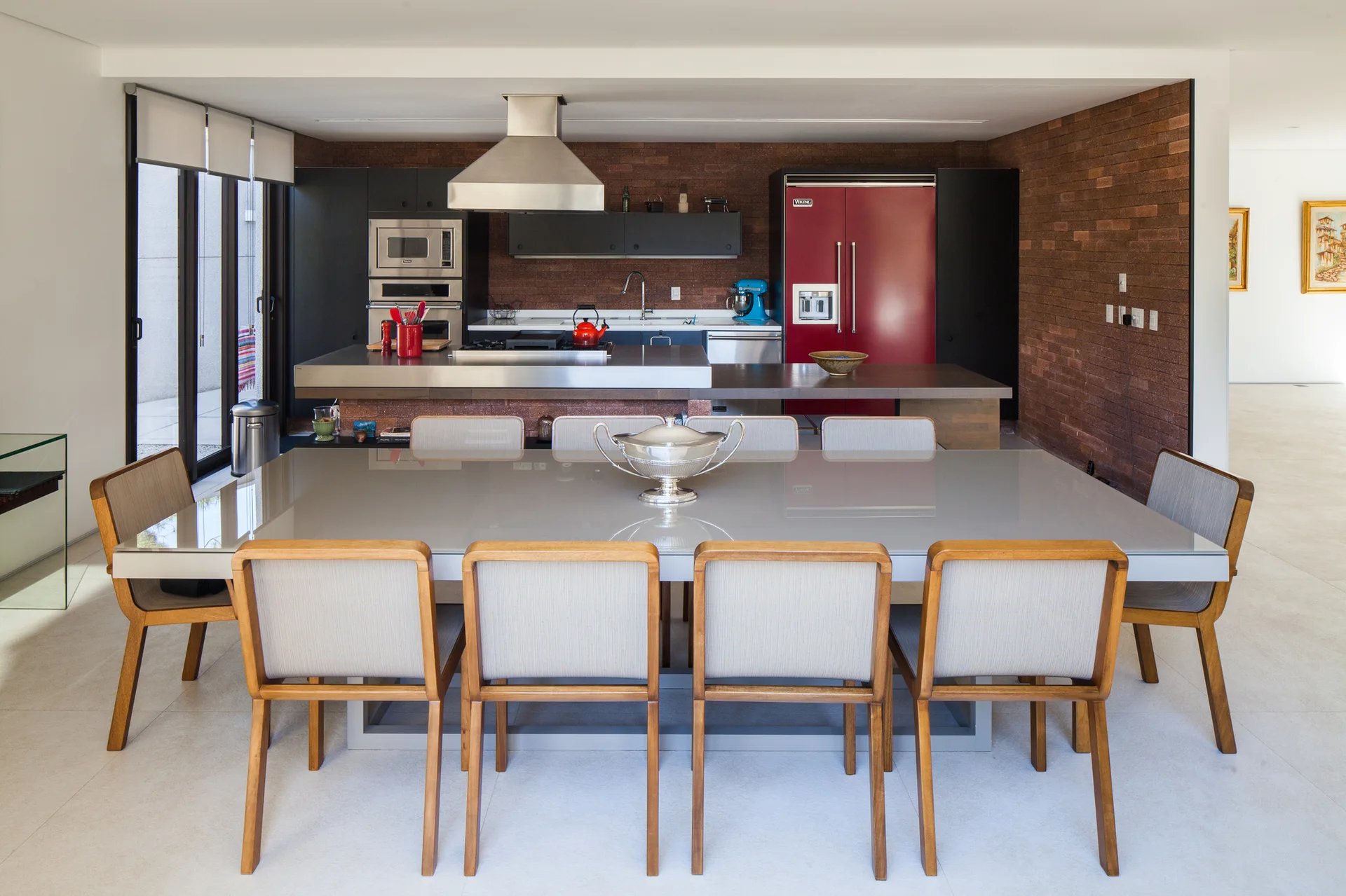 A dining room features a glossy gray table and wooden chairs in front of a kitchen with an exposed brick wall and red refrigerator.