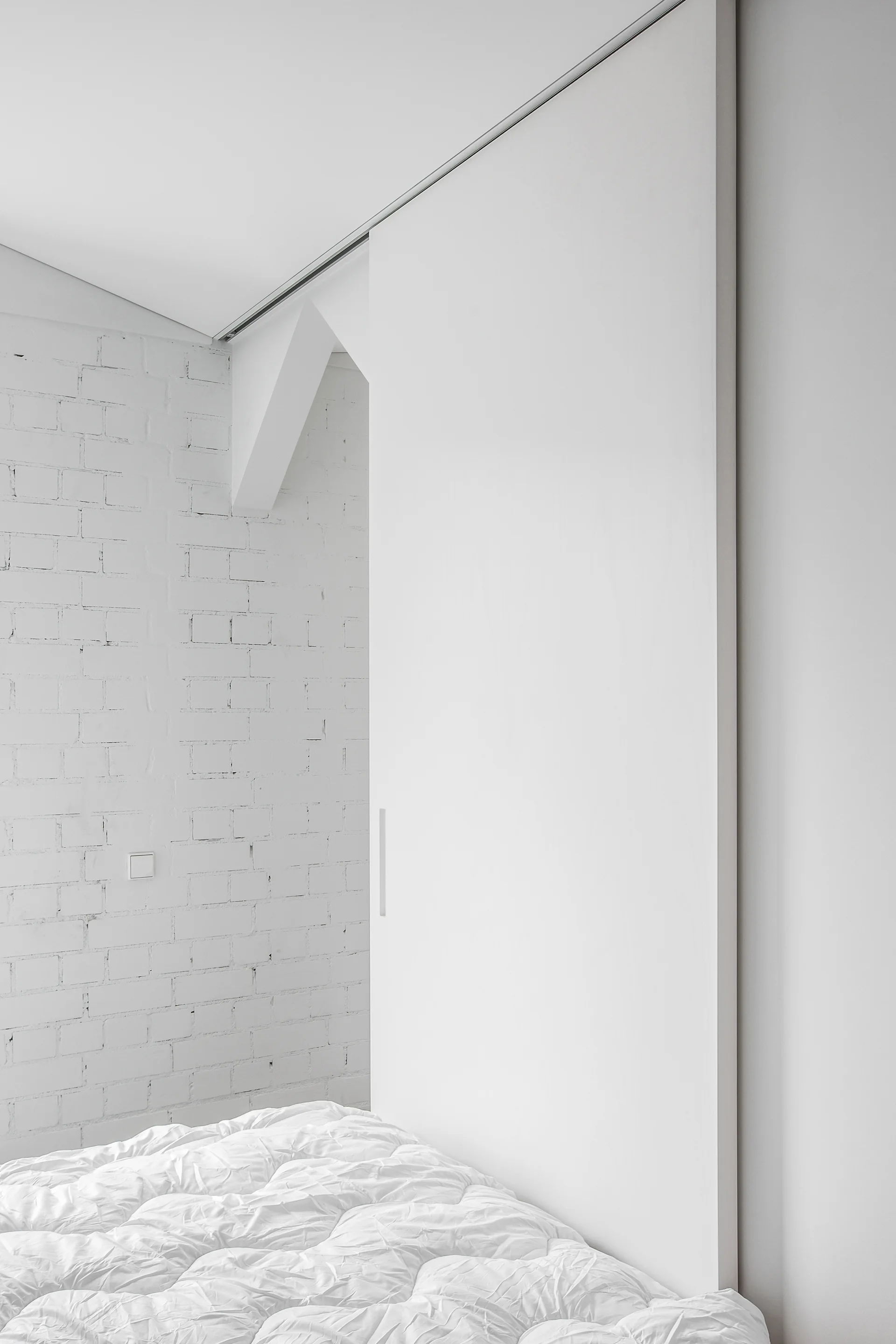 A large white sliding door hangs from a ceiling track next to a painted white brick wall in a minimalist bedroom with textured white bedding.
