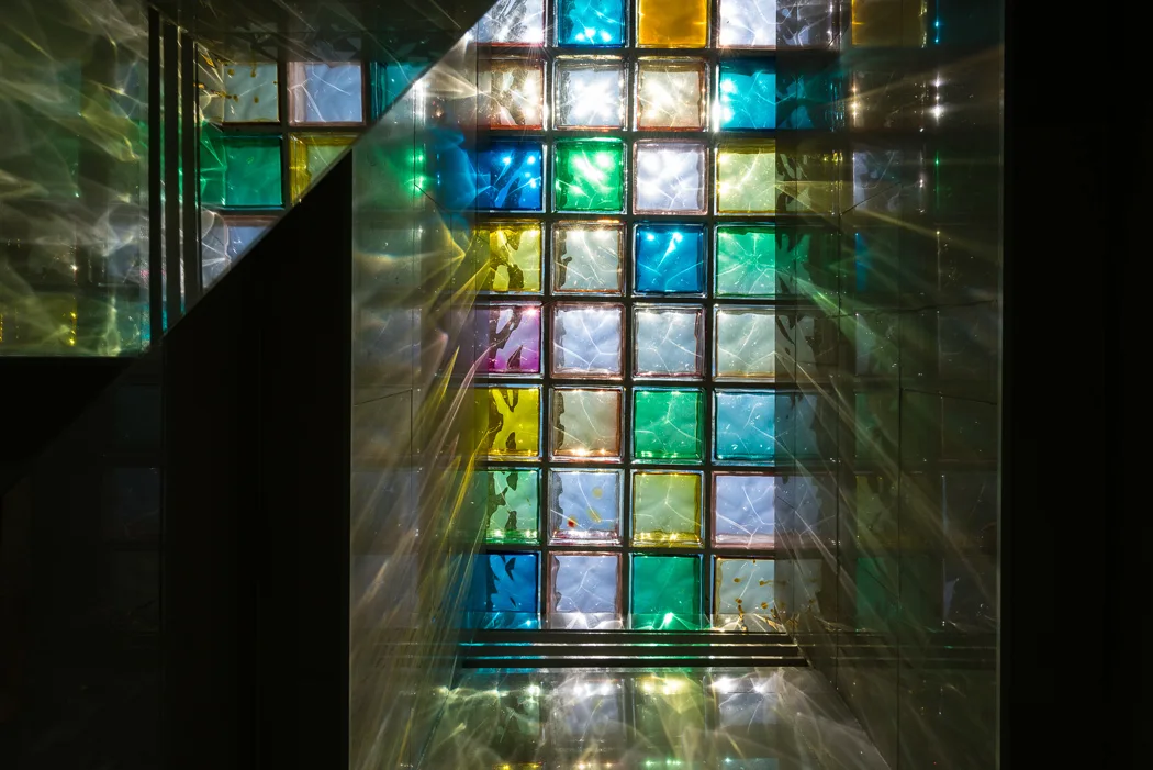 A grid of yellow, green, and blue translucent glass blocks is backlit, casting vibrant reflections onto surrounding dark, glossy surfaces.