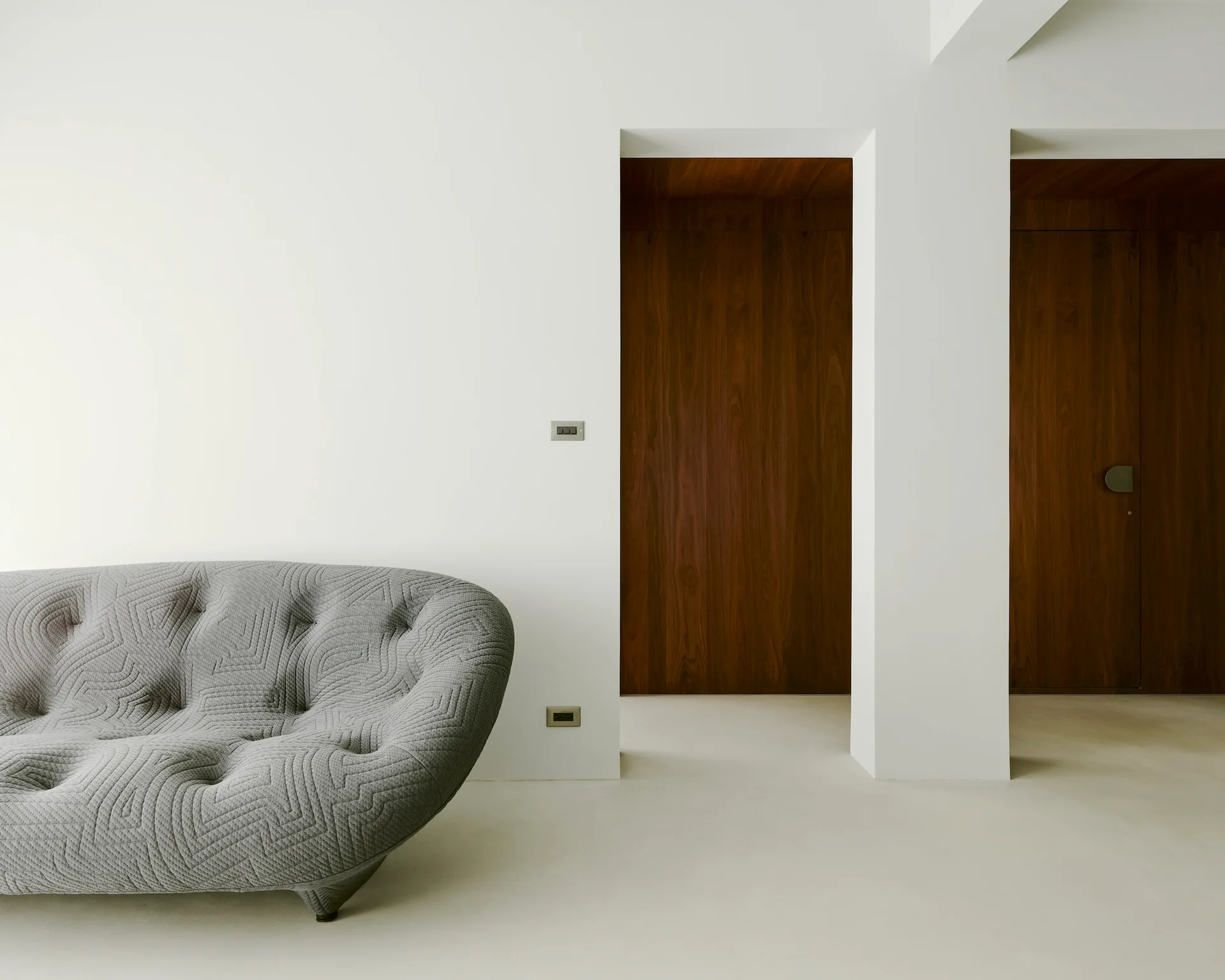 A low-profile tufted gray sofa sits against a white wall next to two recessed dark wood doorways.