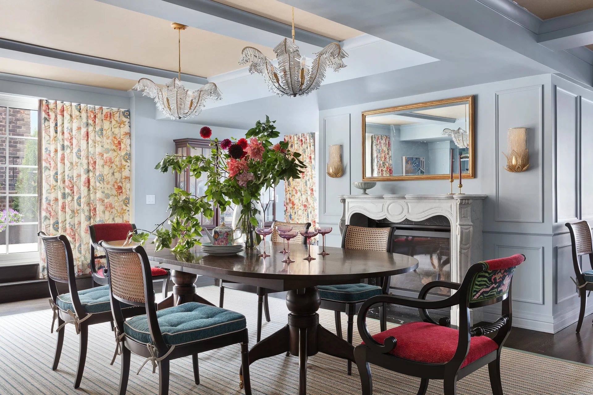 A dining room features a dark wood table surrounded by cane-back chairs on a striped rug, set against light blue paneled walls and a marble fireplace.