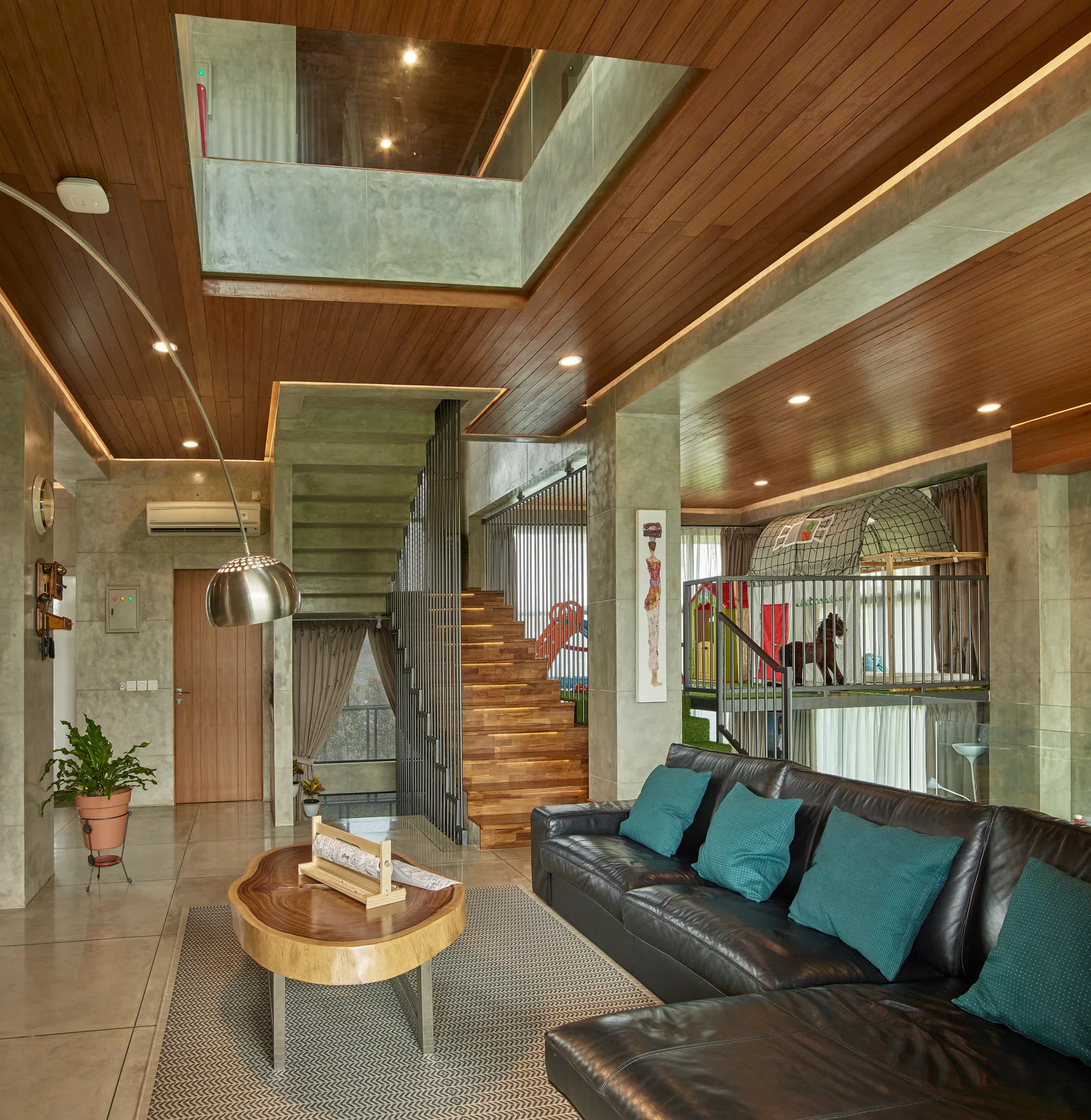 A modern living room featuring a black leather sectional sofa, concrete walls, a warm wood plank ceiling, and an open wooden staircase.