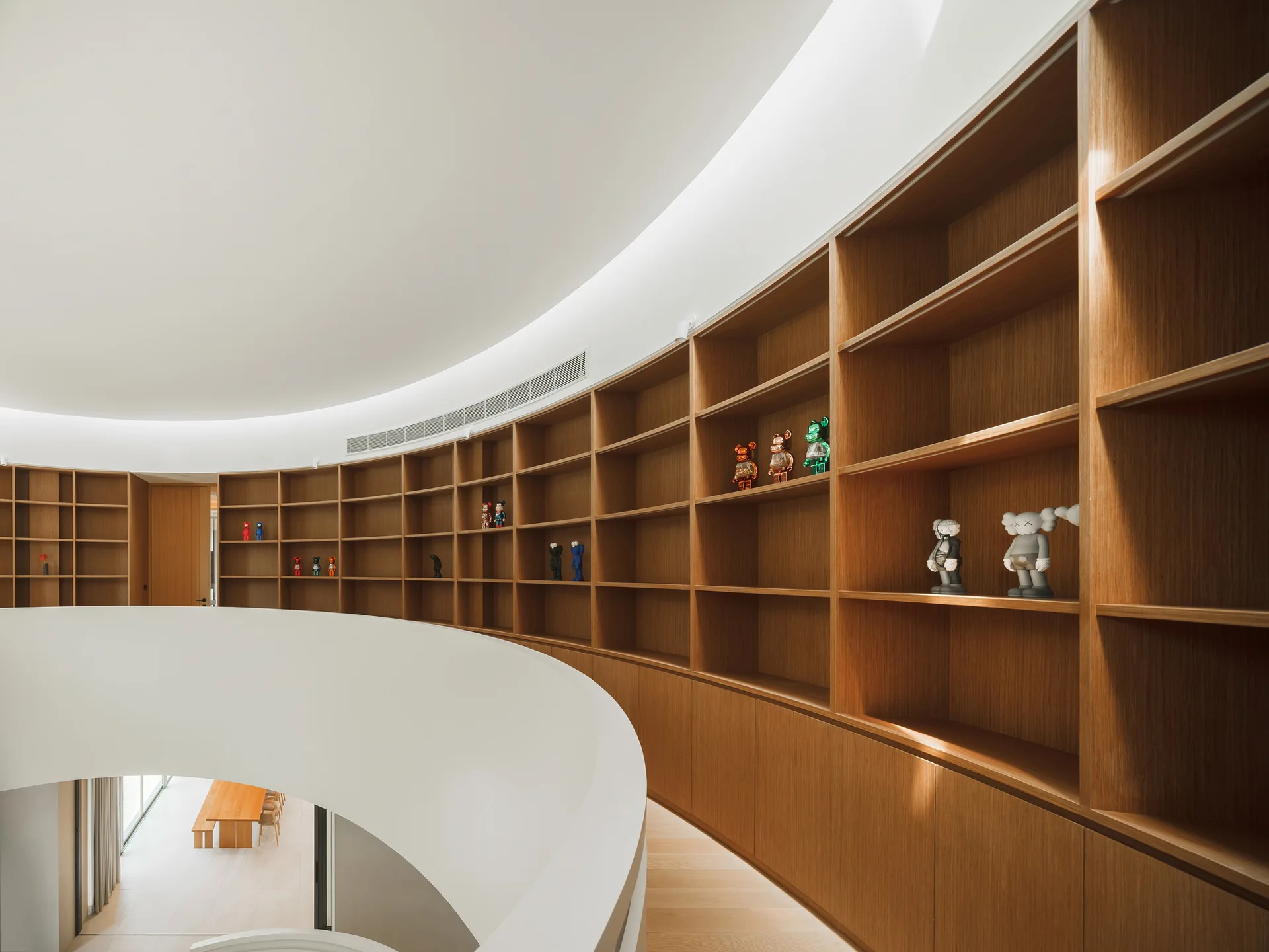 A curved wood built-in bookshelf lines a white landing overlooking a modern atrium with sparse figurines on display.