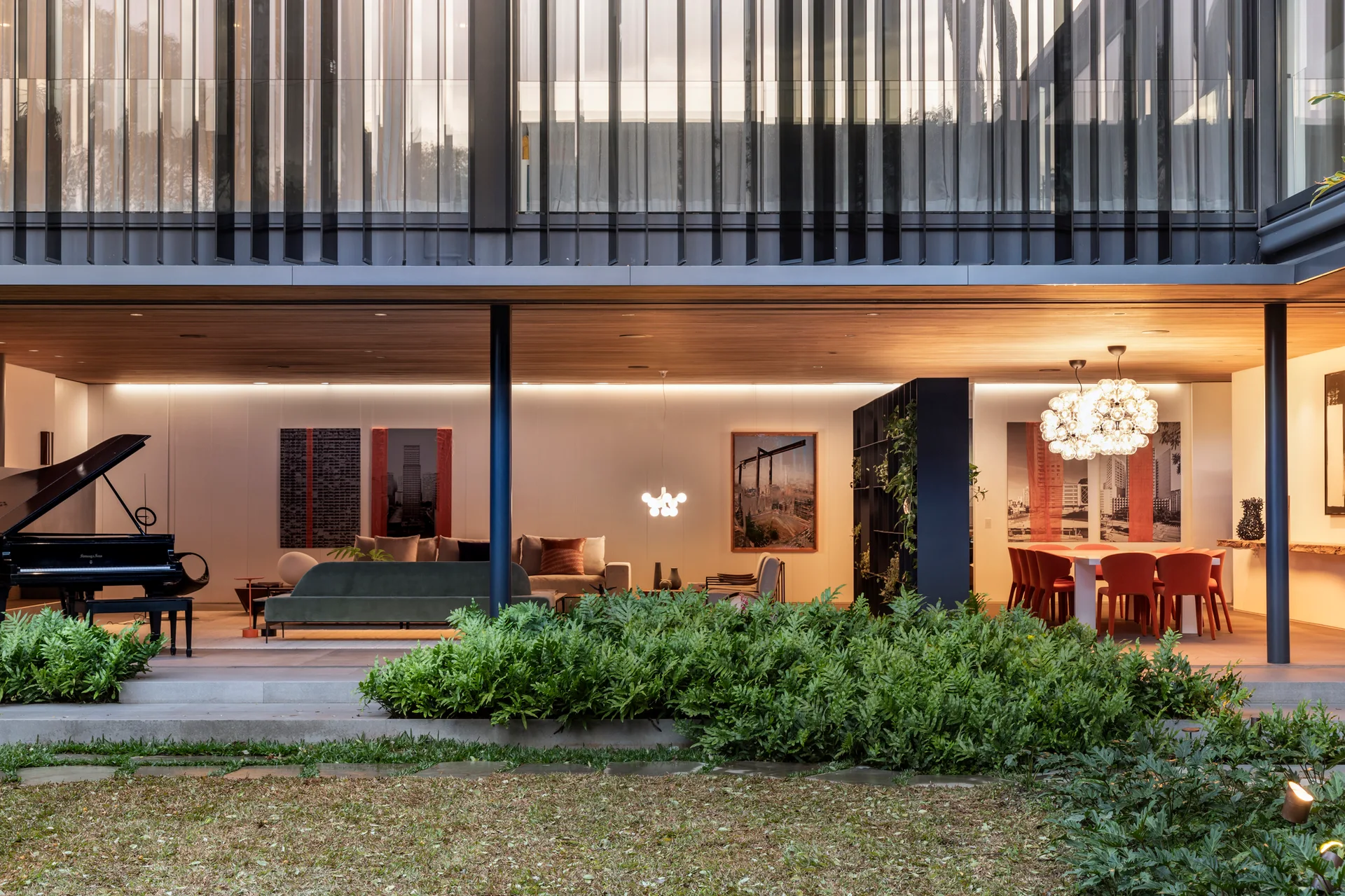 An open-plan living and dining area with a grand piano, green sofa, and orange dining chairs underneath a continuous warm wood ceiling next to a lawn.