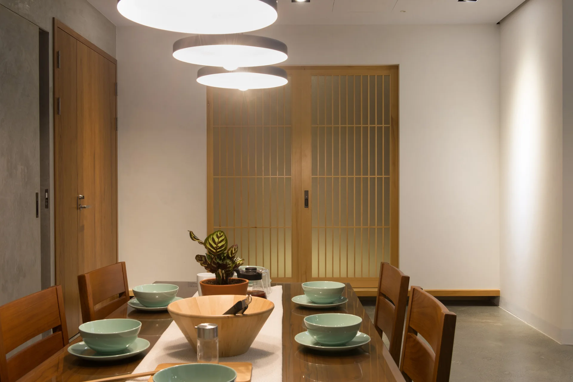 A wooden dining table set with mint green bowls sits under three round pendant lights in front of a shoji-style sliding wood door.