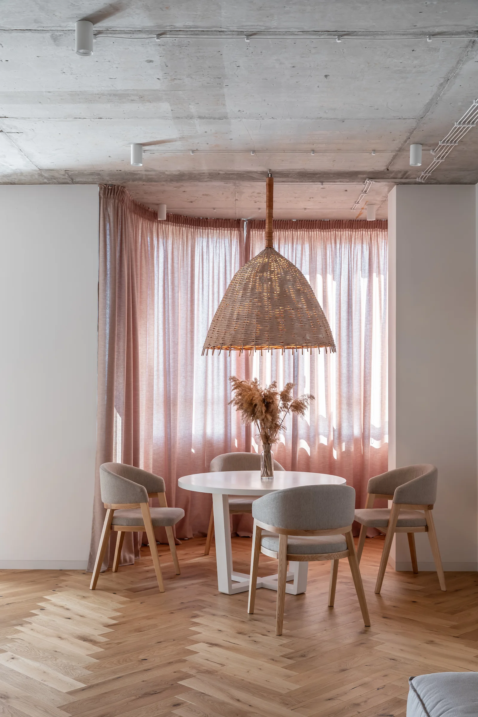 A round dining table and grey chairs sit before floor-length pink curtains under an exposed concrete ceiling and a large woven pendant.