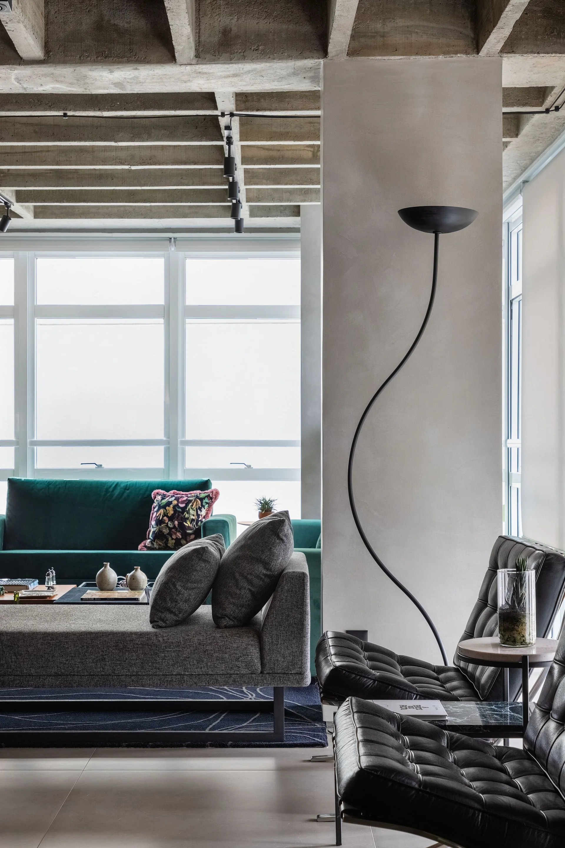 A modern living room with exposed concrete ceiling beams, a central support column, a curved black floor lamp, and tufted black leather chairs.