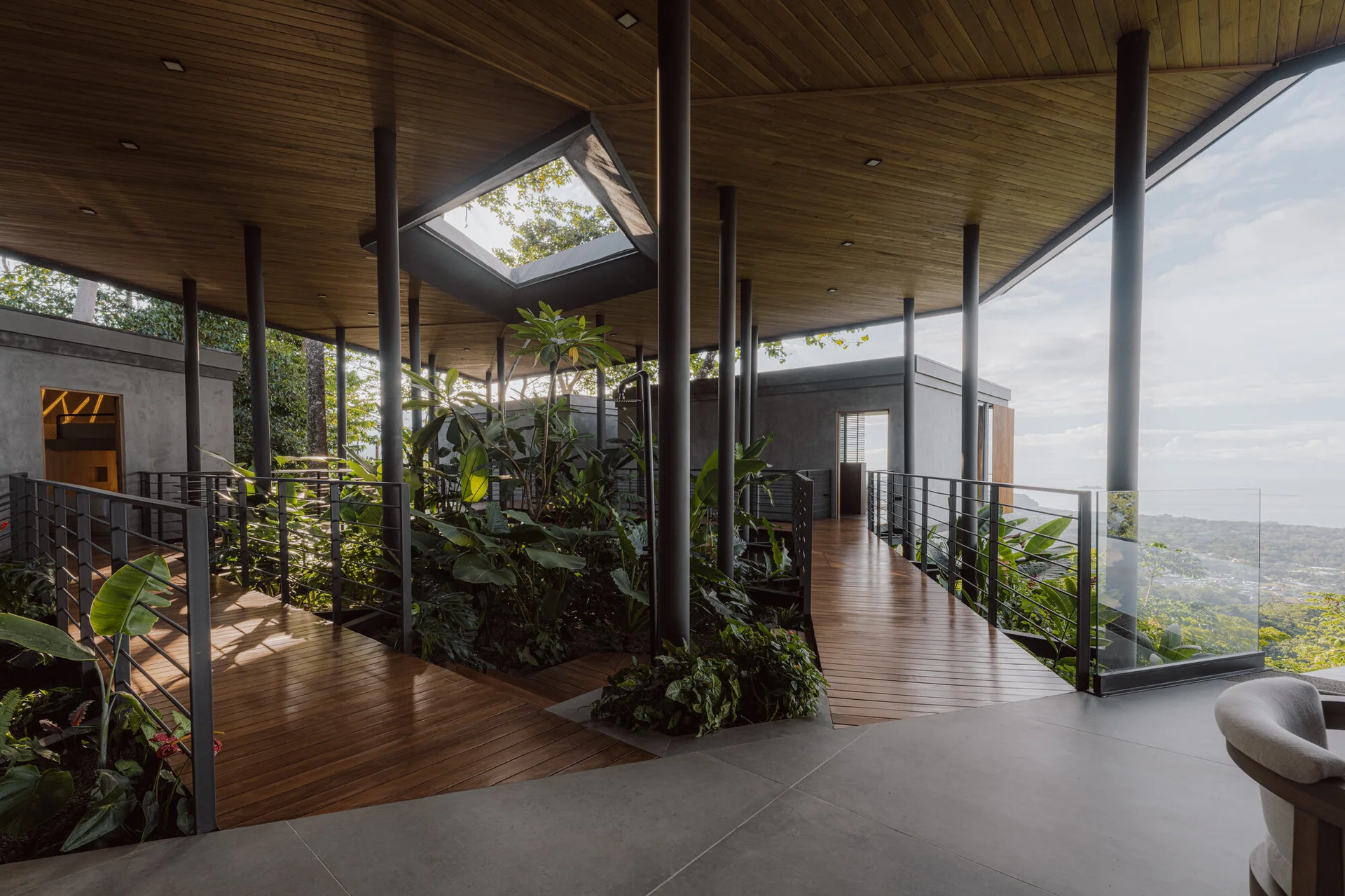 An open-air terrace features wooden walkways, slender black columns supporting a timber ceiling, and lush green plants beneath a square skylight.