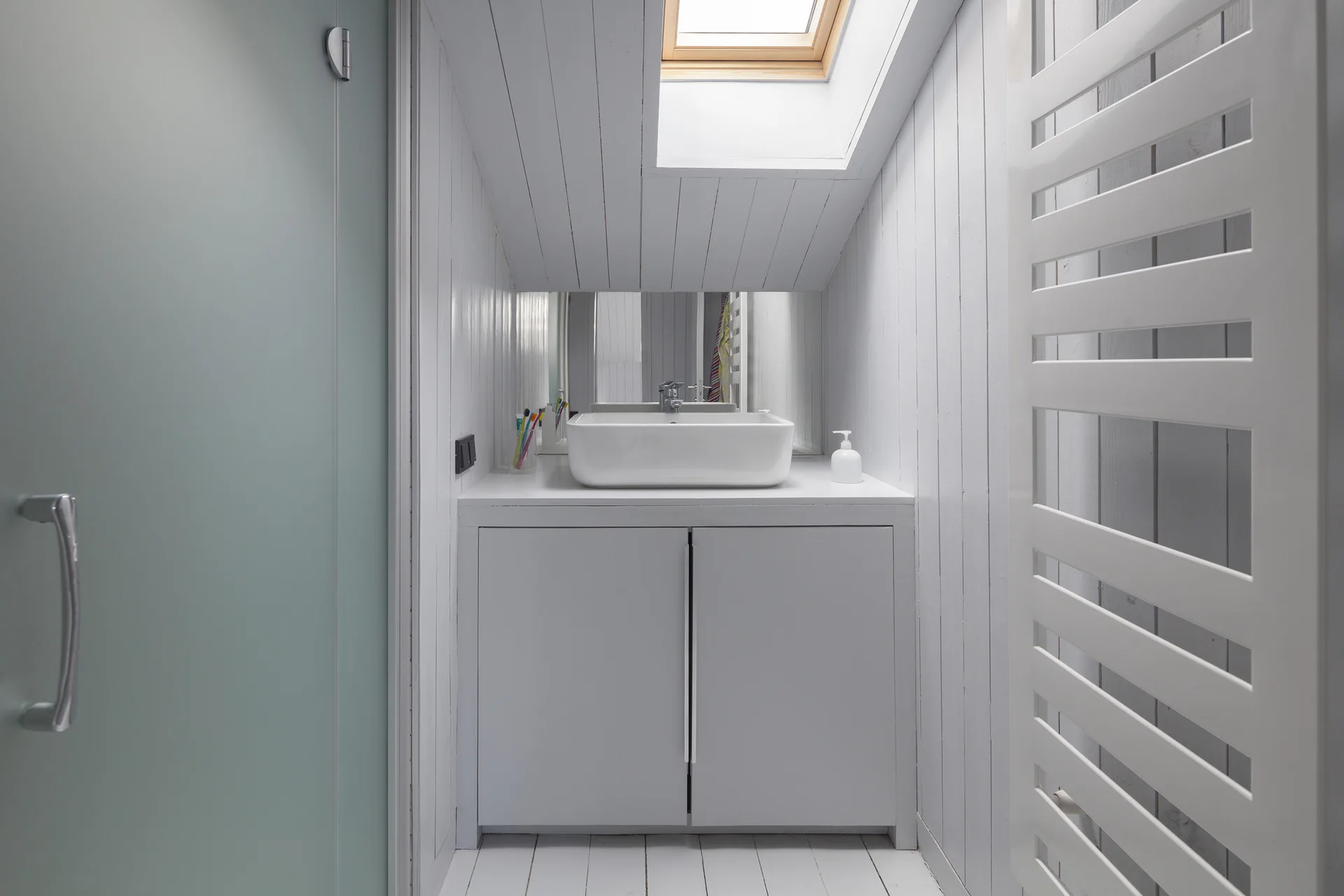 A white attic bathroom features a vessel sink vanity, vertical wood-paneled walls, and a sloped ceiling with a bright skylight.