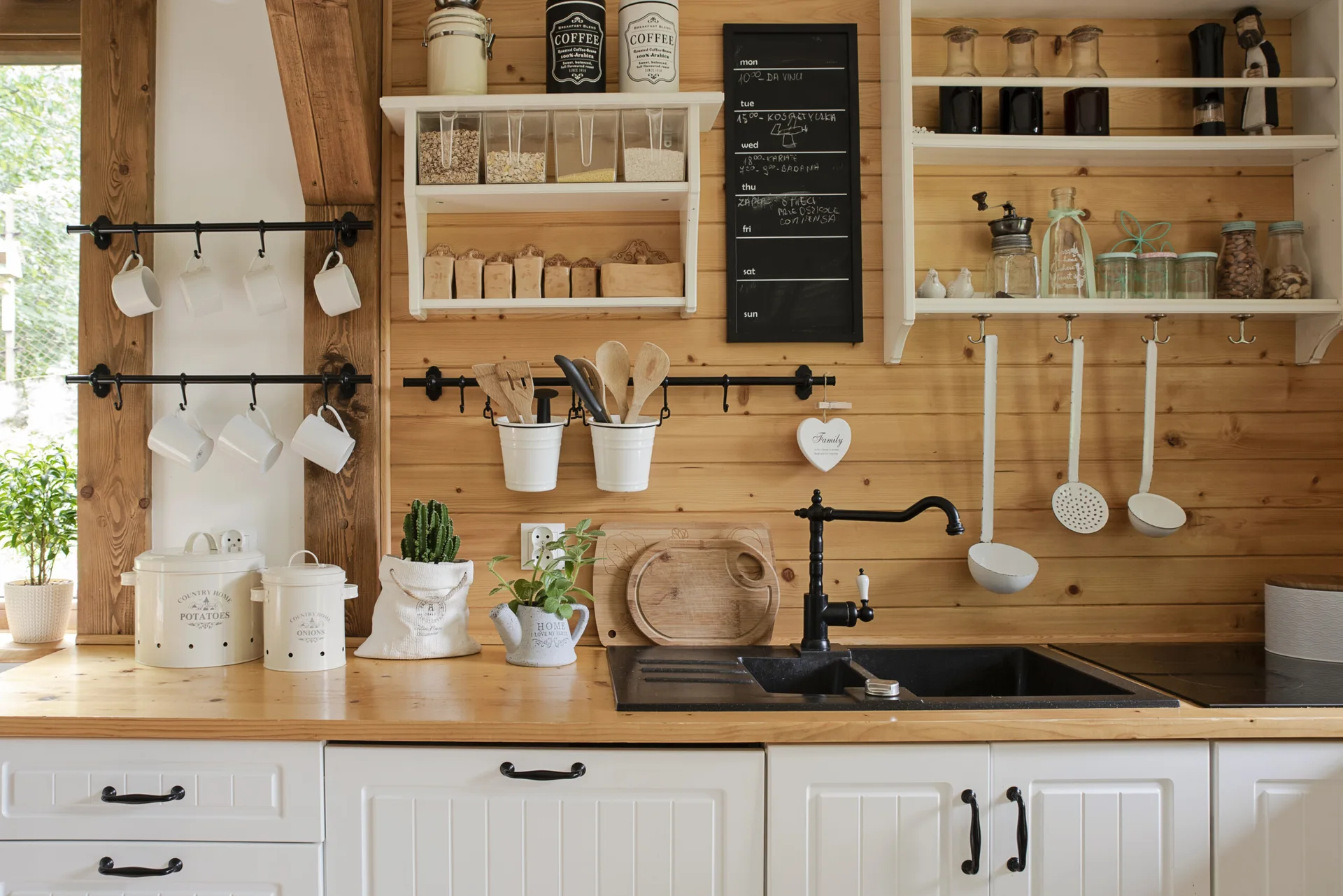 A farmhouse kitchen features horizontal wood panel walls, white shaker cabinets, and a black sink with matching matte black rails holding white mugs.
