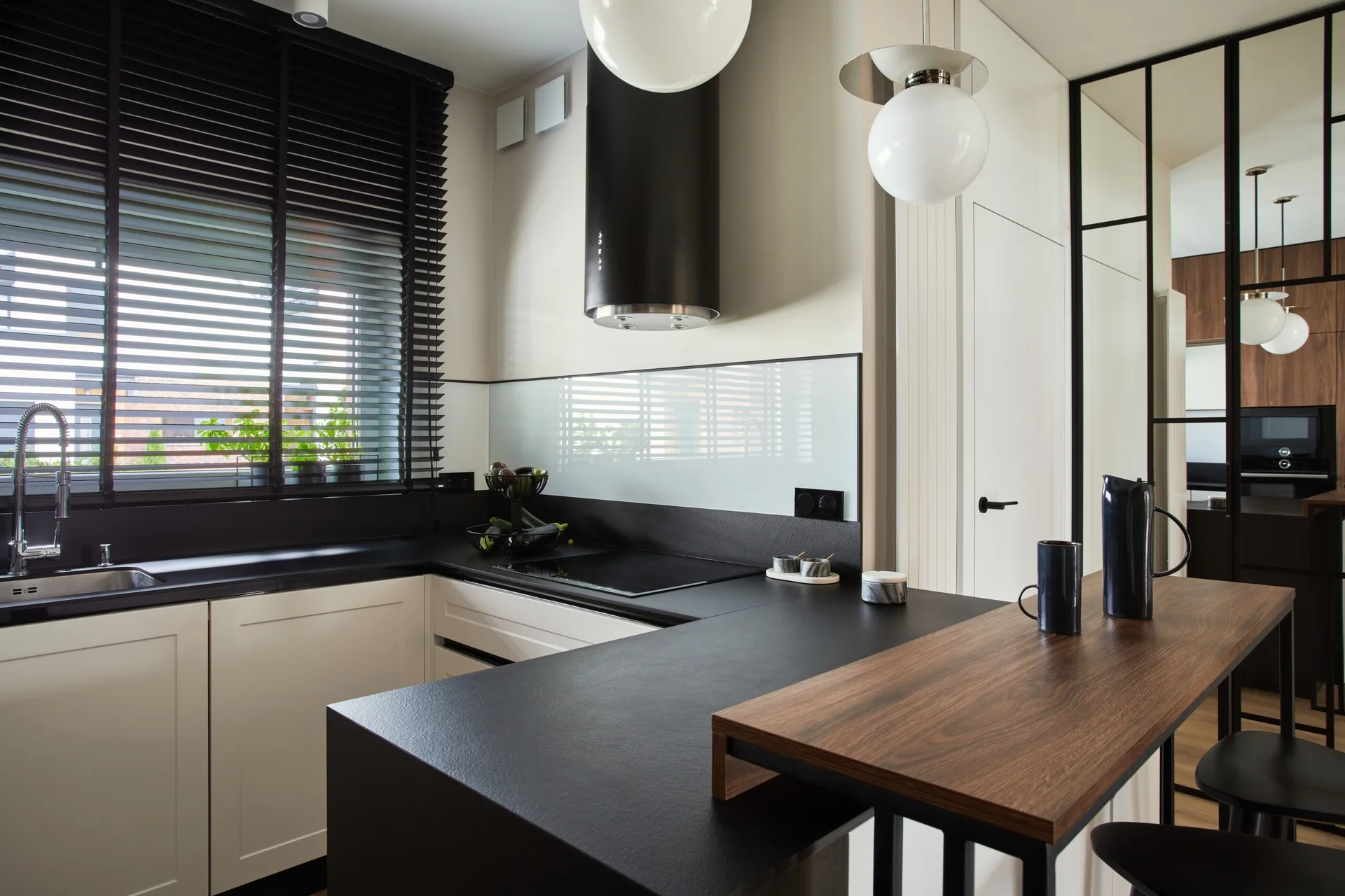 A modern kitchen featuring cream cabinets, black countertops, a cylindrical range hood, black blinds, and a raised wooden breakfast bar.