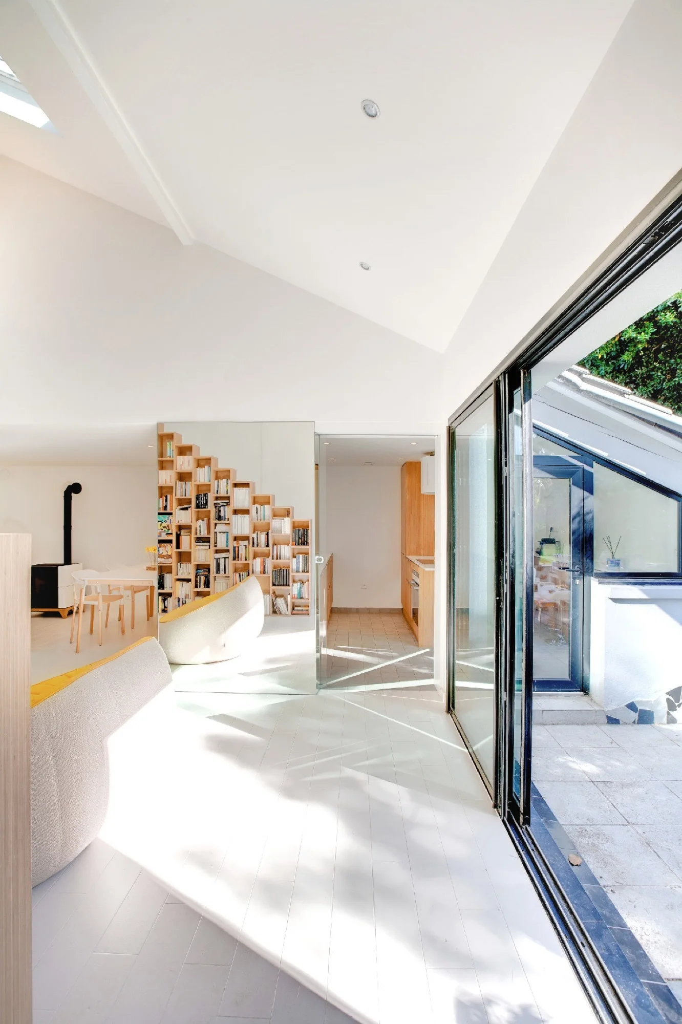 A bright Scandinavian living room featuring a stepped wooden bookshelf, a mirrored accent wall, and large sliding glass doors leading to a patio.
