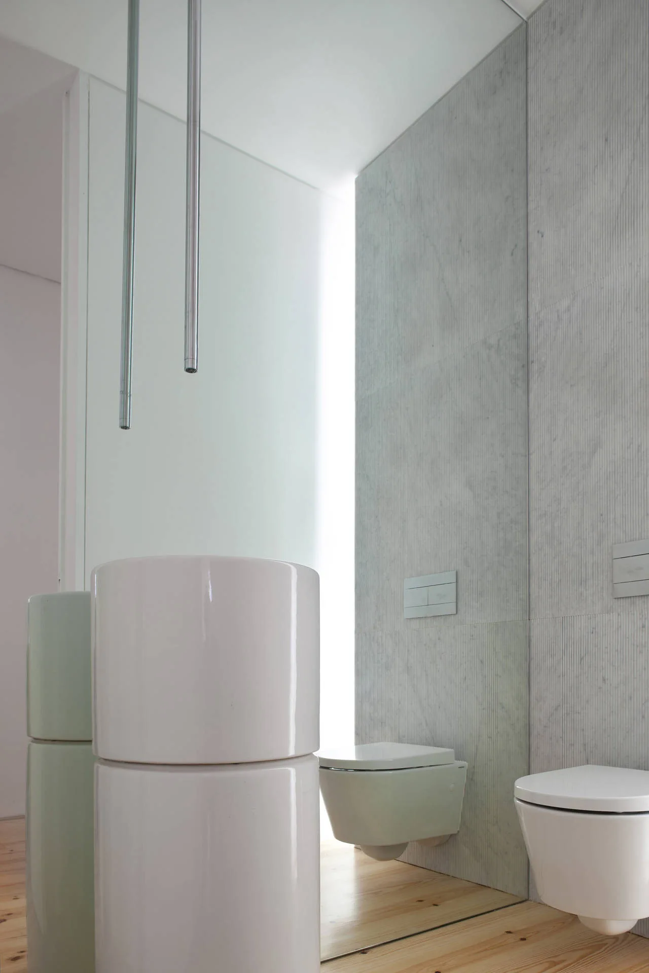 A minimalist bathroom with vertical fluted stone wall panels, a wall-mounted toilet, a white cylindrical sink, and slim ceiling-mounted metallic faucets.