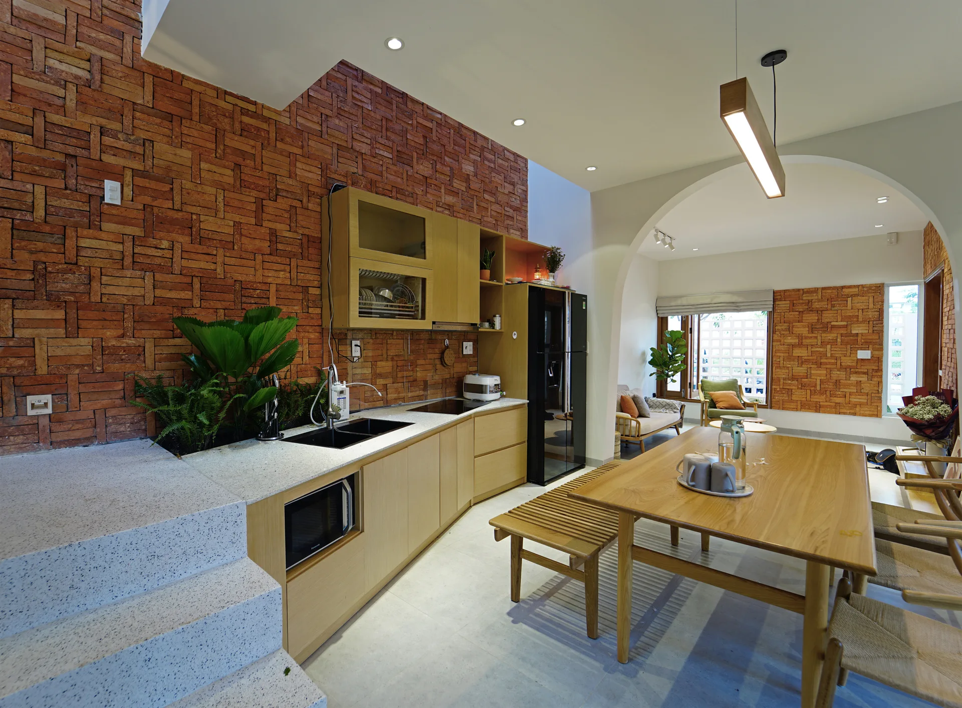 A modern kitchen featuring an exposed red brick wall, light wood cabinetry, a wooden dining table with a bench, and a curved plaster archway.