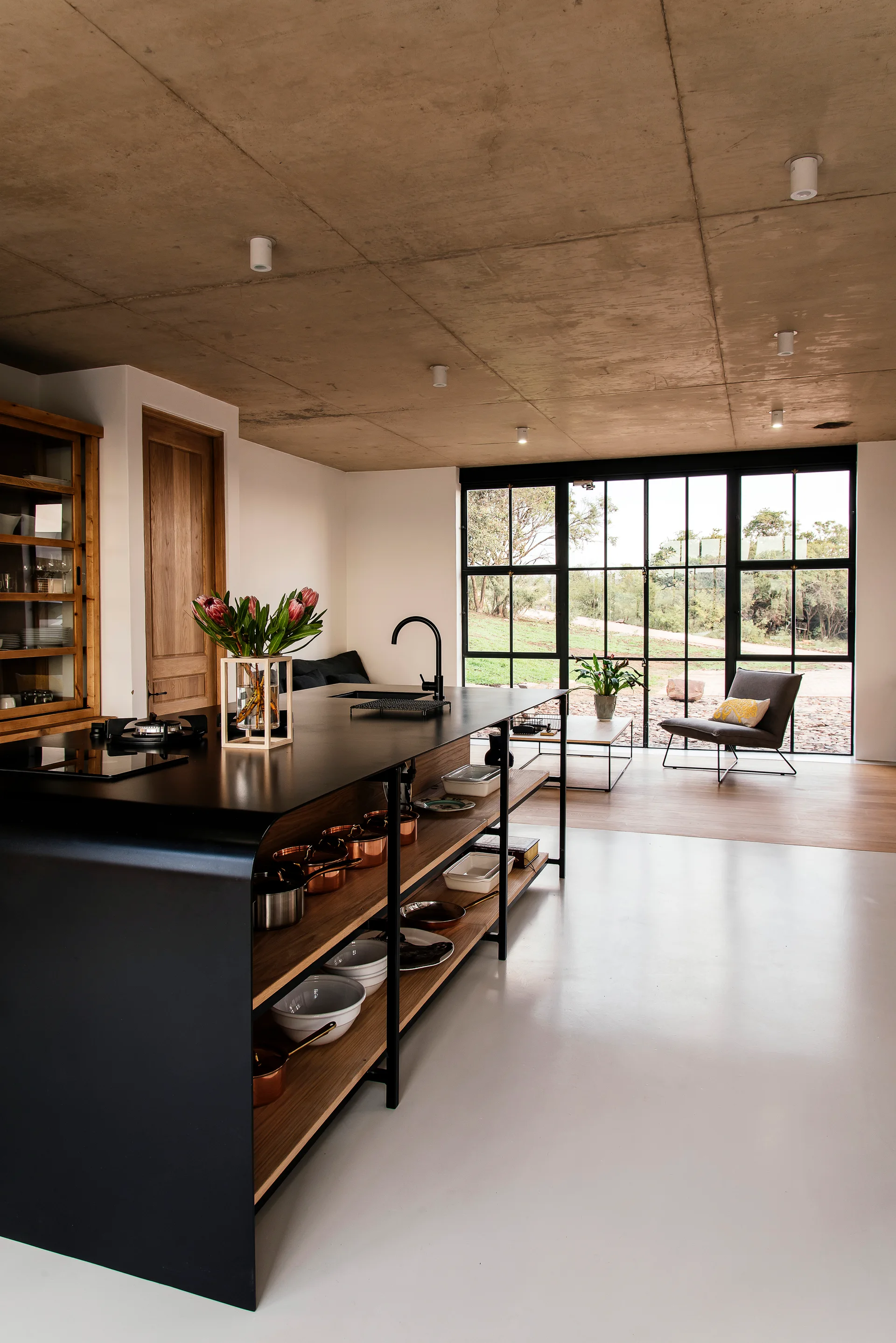 A modern kitchen featuring a black island with open wood shelving, an exposed concrete ceiling, and a large black-framed glass wall leading outside.