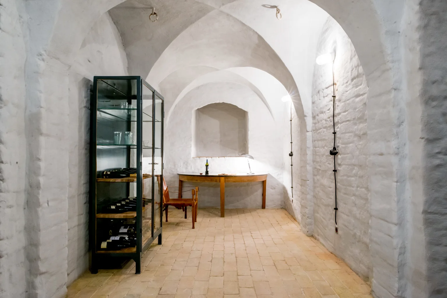 A white vaulted brick wine cellar features a dark glass-front display cabinet, a wooden console table, and tan brick flooring.