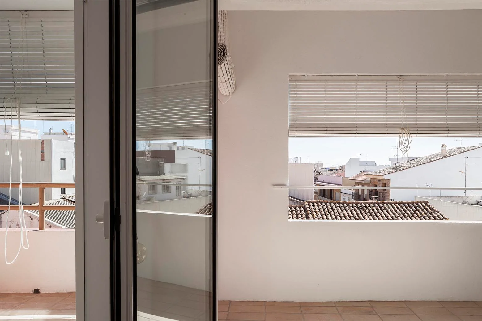 A sunlit Mediterranean terrace featuring white plaster walls, terracotta floor tiles, a sliding glass door, and horizontal blinds framing a city rooftop view.