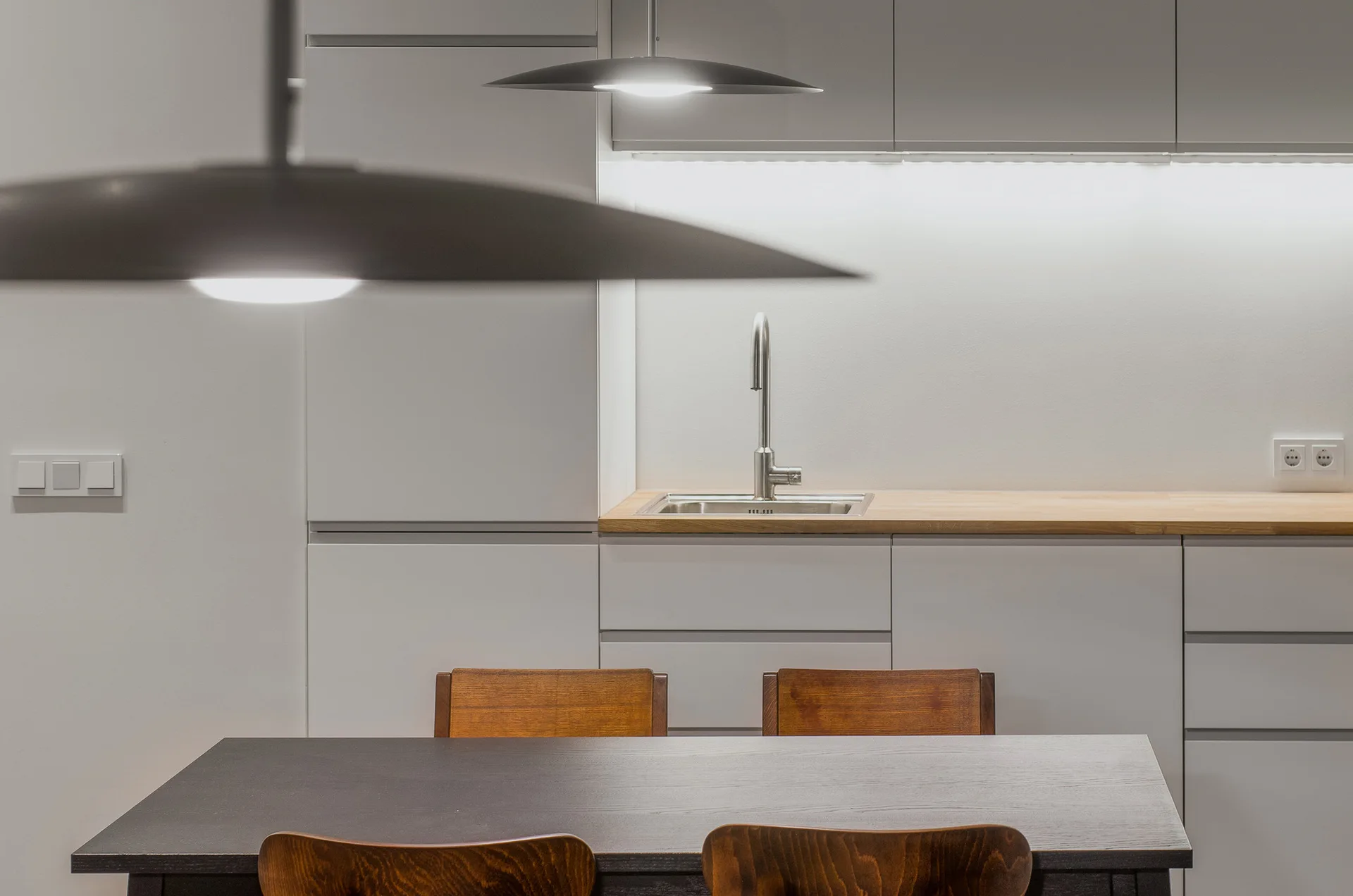 A minimalist grey kitchen featuring handleless cabinets, a light wood countertop with a stainless steel sink, and a dark wood dining table in the foreground.