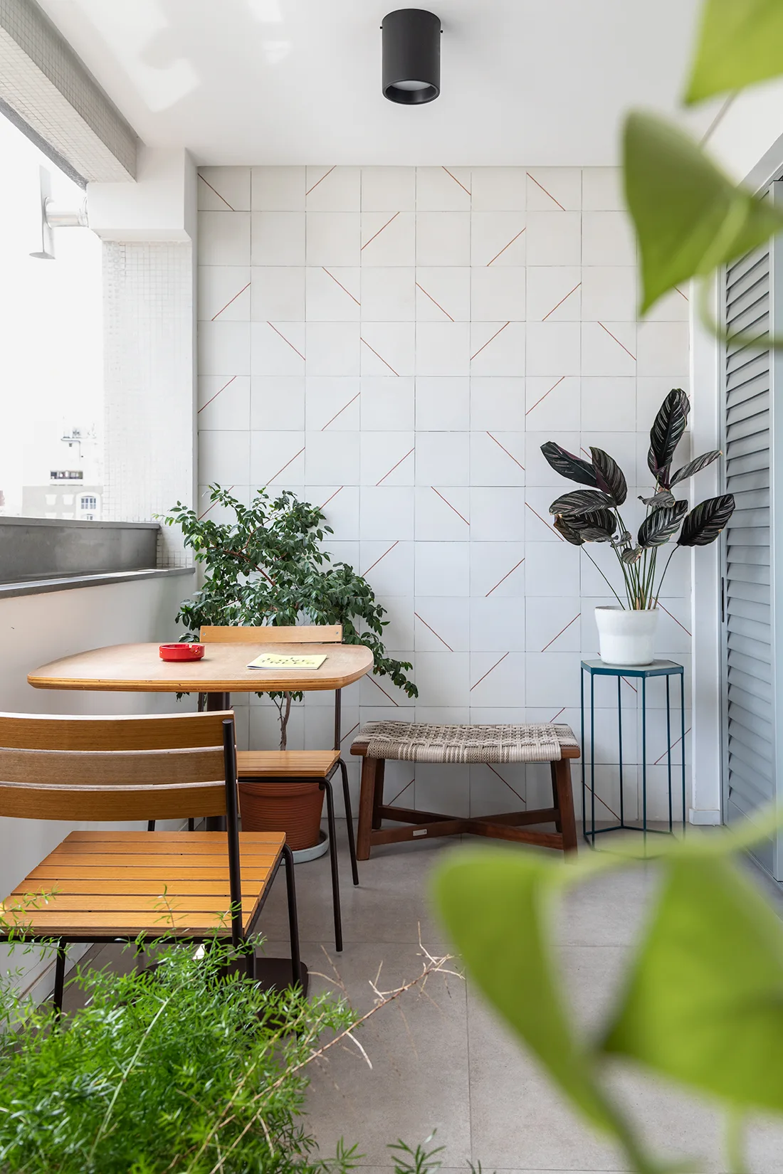 A bright apartment balcony features a wooden dining table and chairs, a woven bench, and potted plants against a white wall with geometric tile patterns.