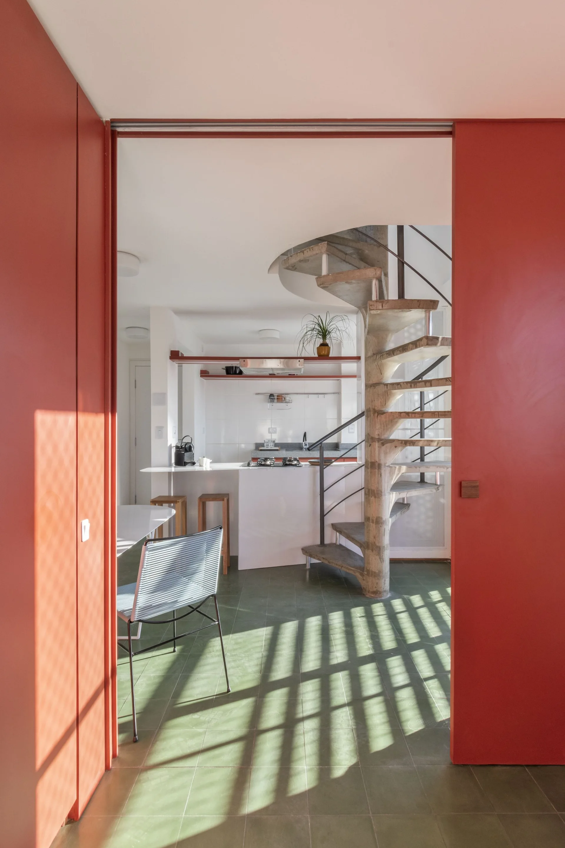 A view through a bright red doorway reveals a kitchen with green floor tiles and a prominent concrete spiral staircase.