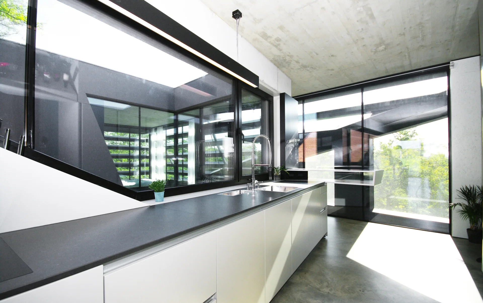 A modern minimalist kitchen featuring white handleless cabinets, a dark countertop, large black-framed windows, and an exposed raw concrete ceiling.