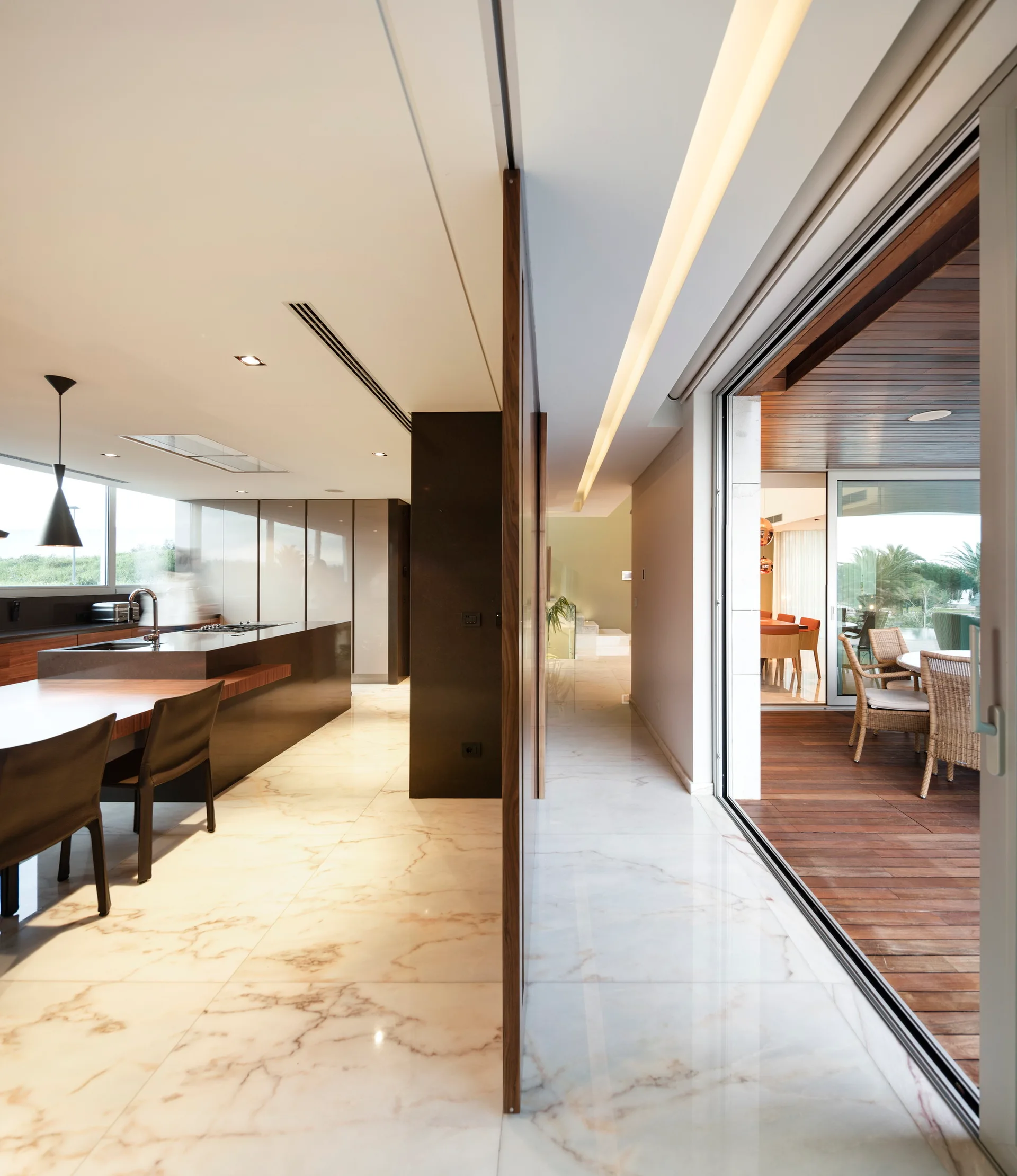 A minimalist kitchen with polished marble floors features a dark island and opens through large glass sliding doors to a wood-decked terrace.