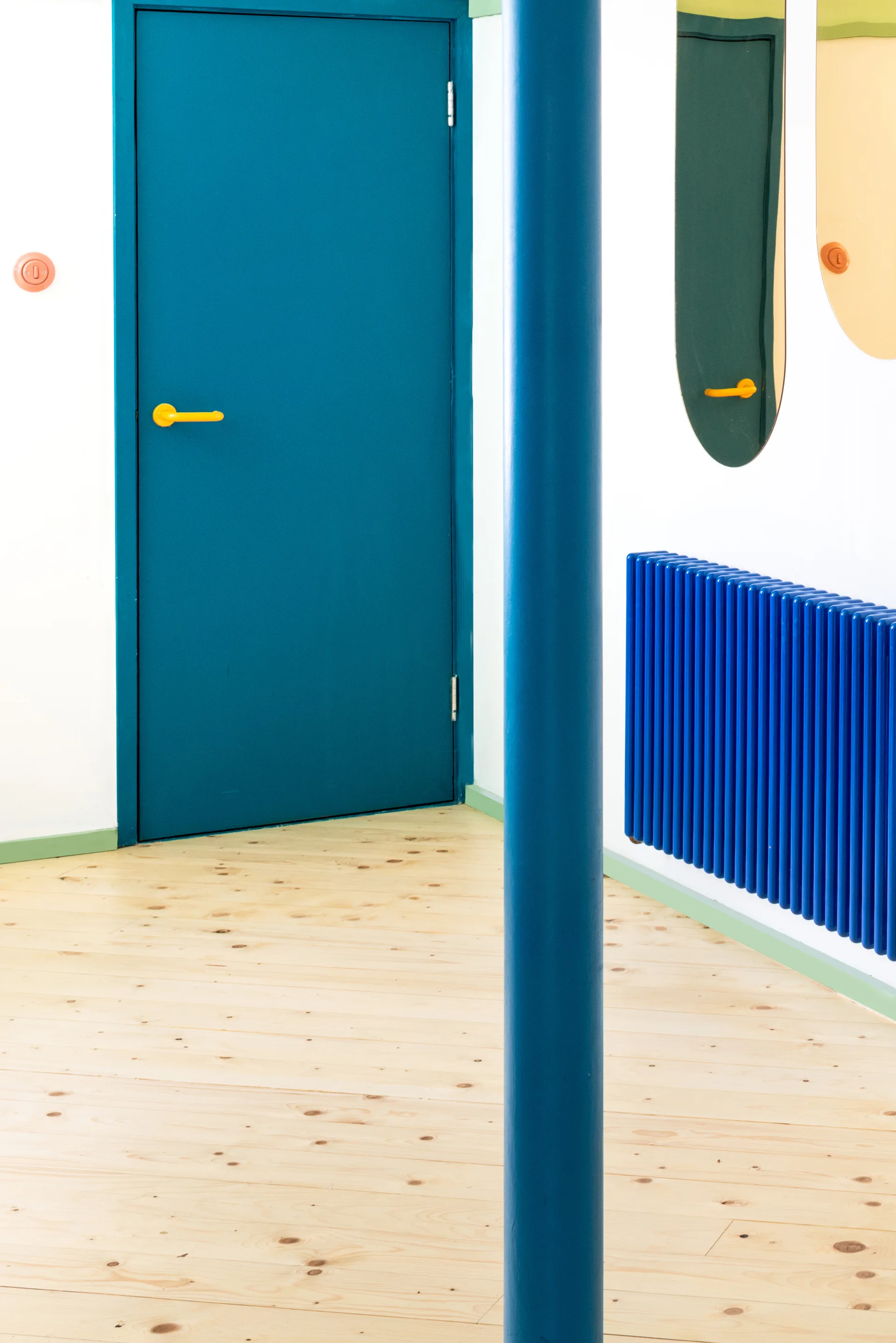 A brightly lit hallway features a bold blue door with a yellow handle, a matching blue structural column, a blue radiator, and light wood flooring.