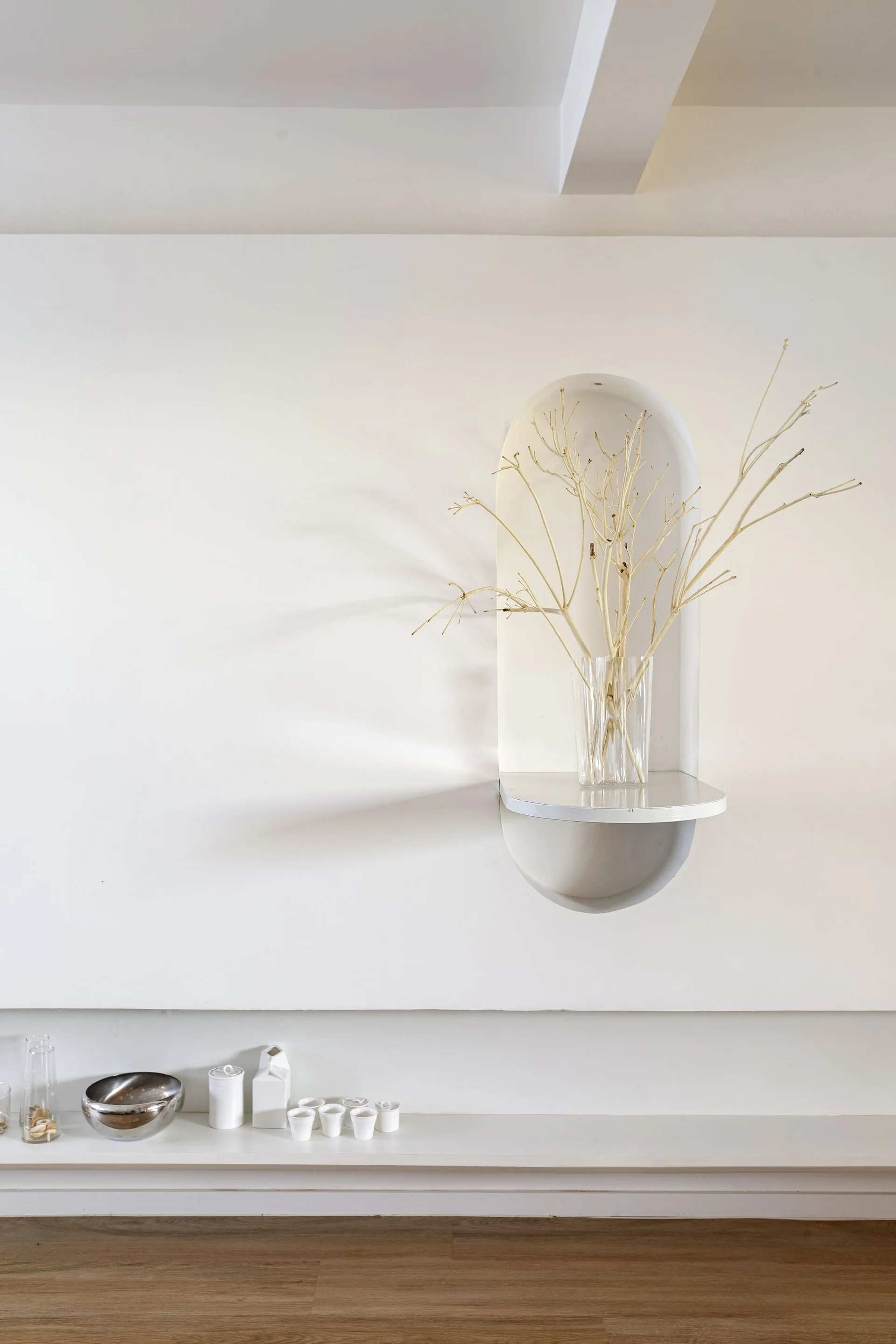 A white bathroom wall features a recessed arched niche holding a glass vase with dried branches above a low shelf with small vessels.