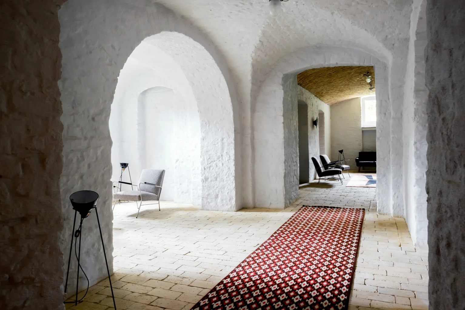 Spacious interior with whitewashed stone archways, tan brick flooring, a patterned red runner rug, and minimalist grey metal-framed armchairs.