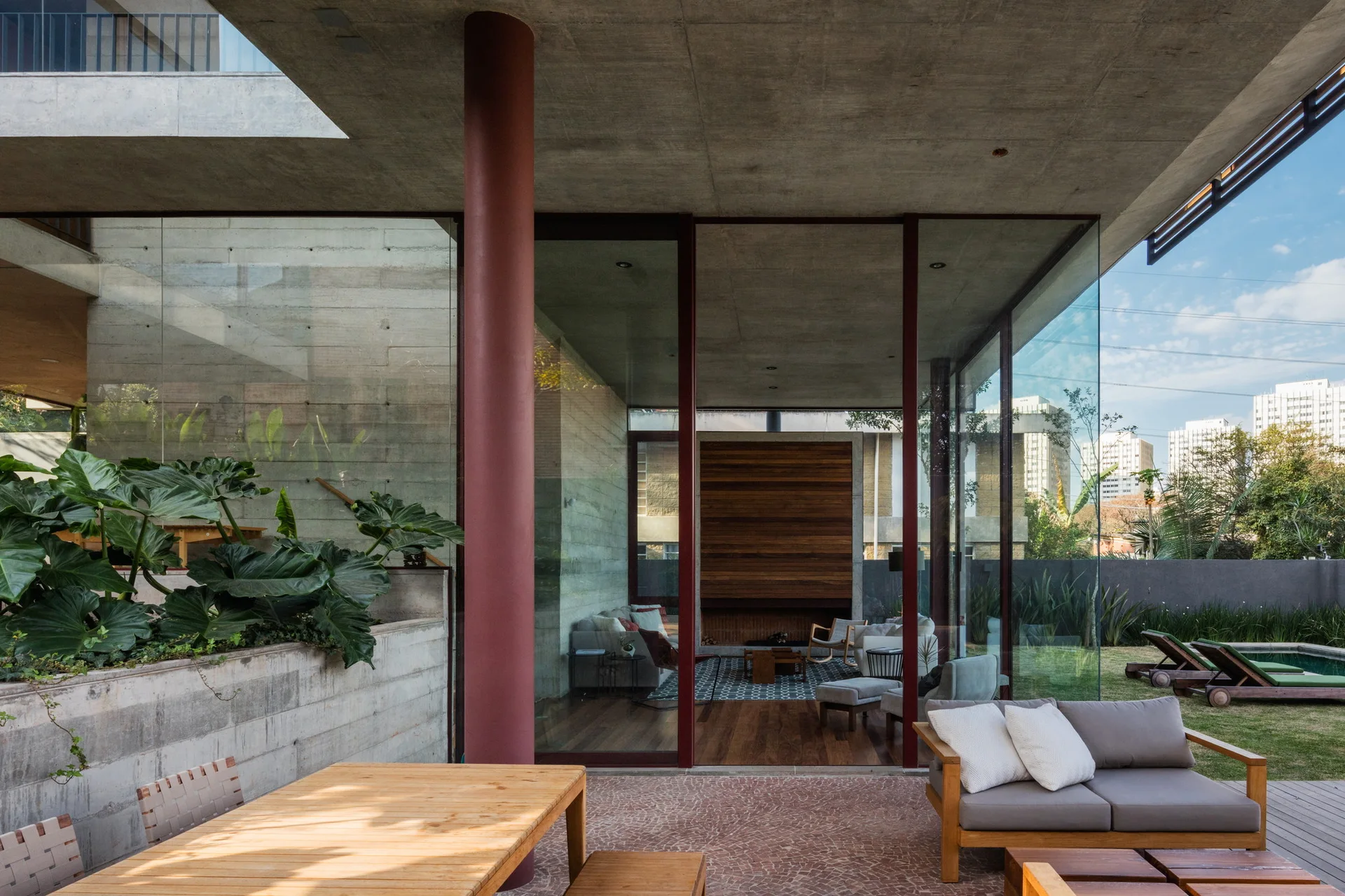 A modern patio with a wood dining table and sofa, separated from an indoor living area by floor-to-ceiling glass walls under a concrete roof.