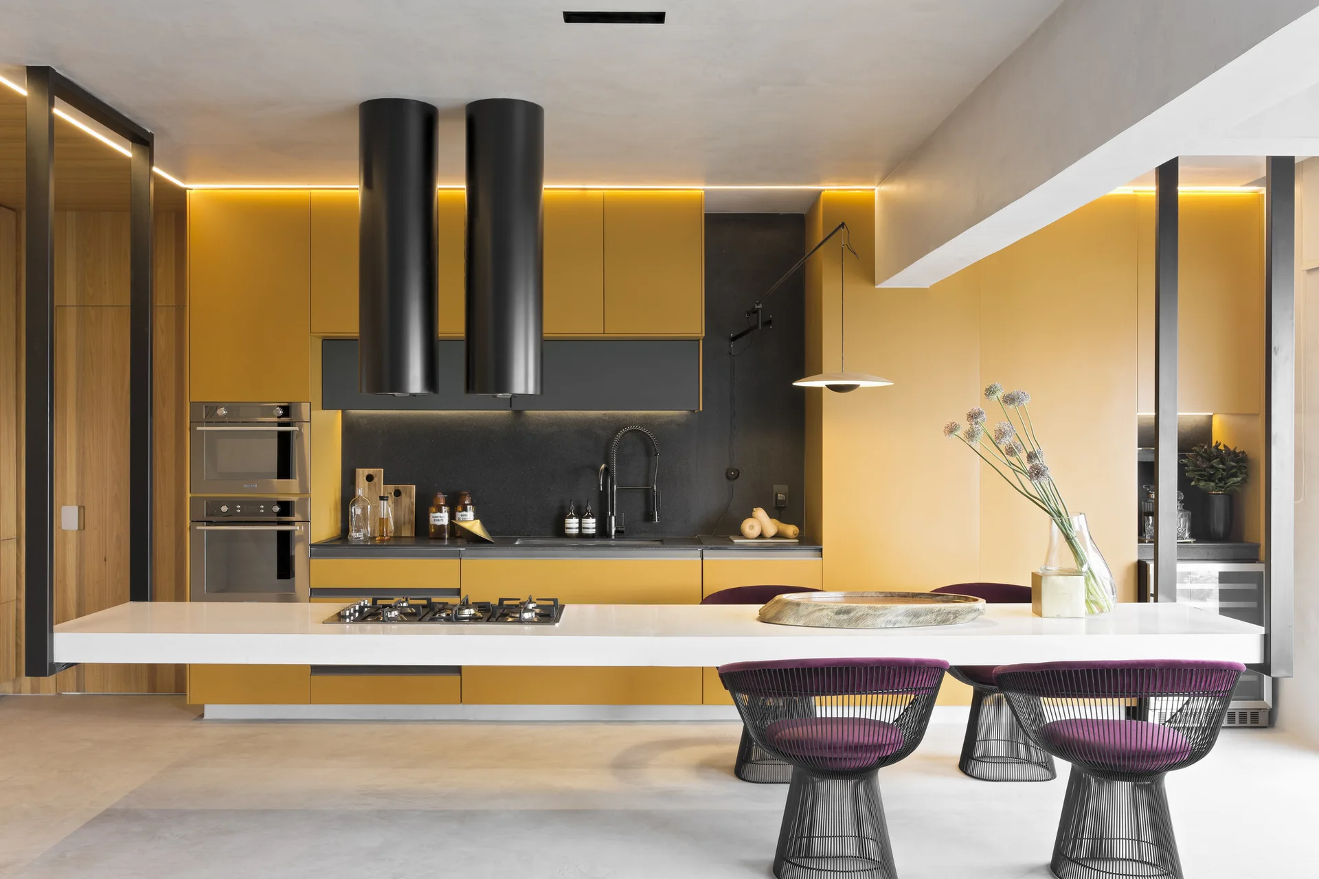A minimalist kitchen features bright yellow cabinets, a long white island with purple wire-frame chairs, and dual cylindrical black range hoods.