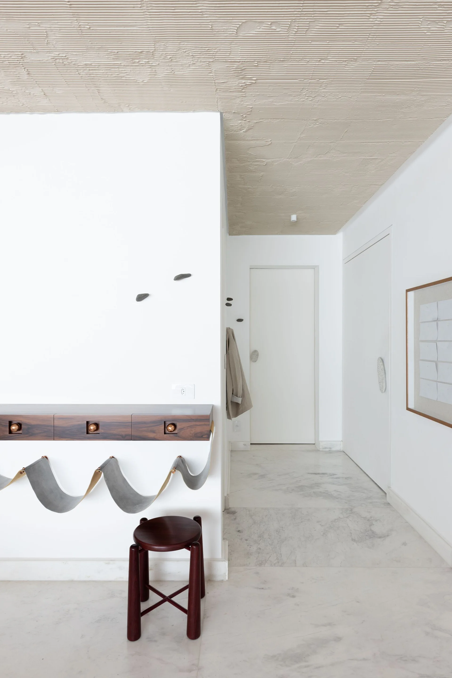 A minimalist entryway features a wall-mounted dark wood console with grey fabric slings, a matching wood stool, and a textured beige ceiling.