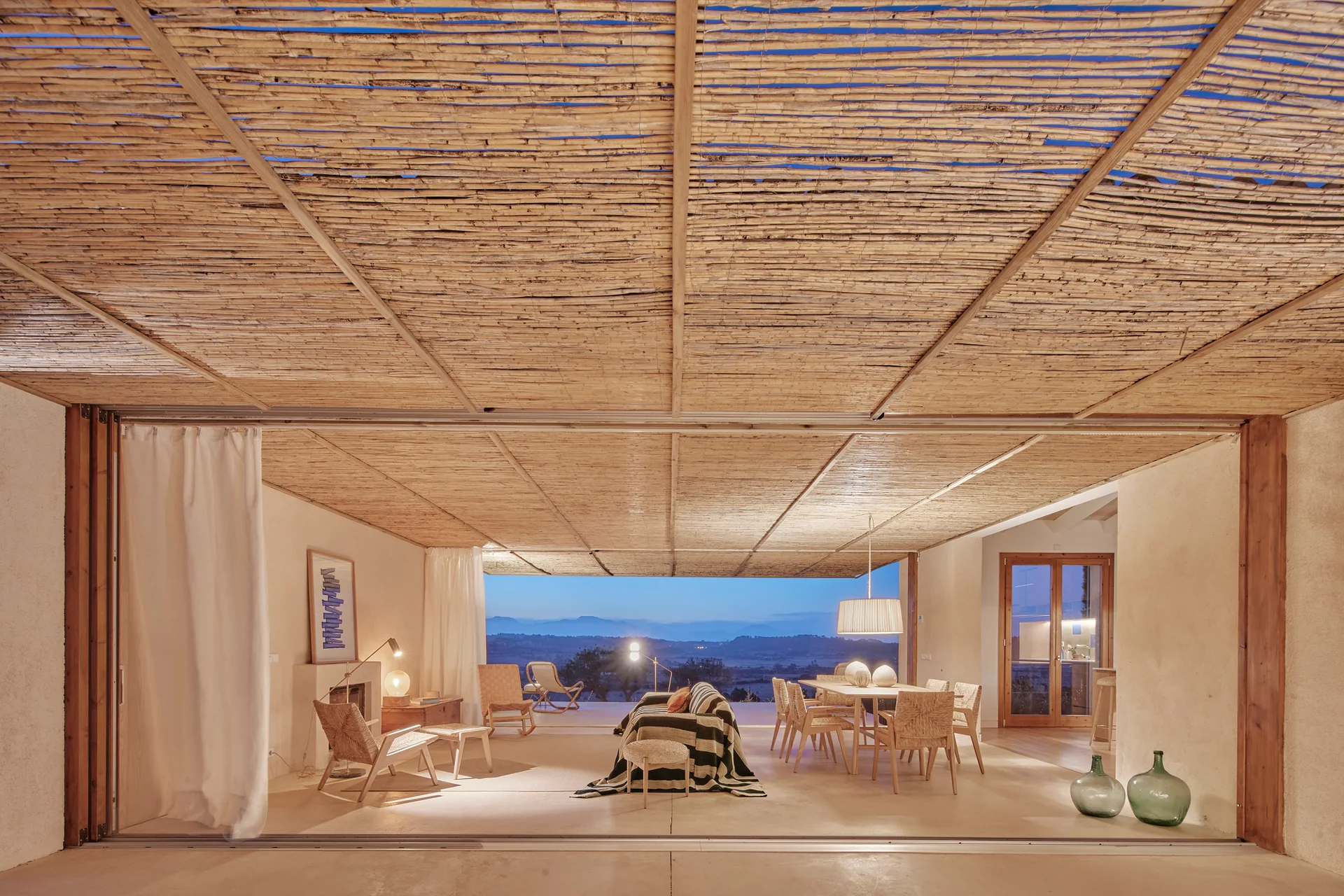 A covered Mediterranean patio featuring a woven bamboo ceiling, a dining set, lounge furniture, and an open view of hills at dusk.