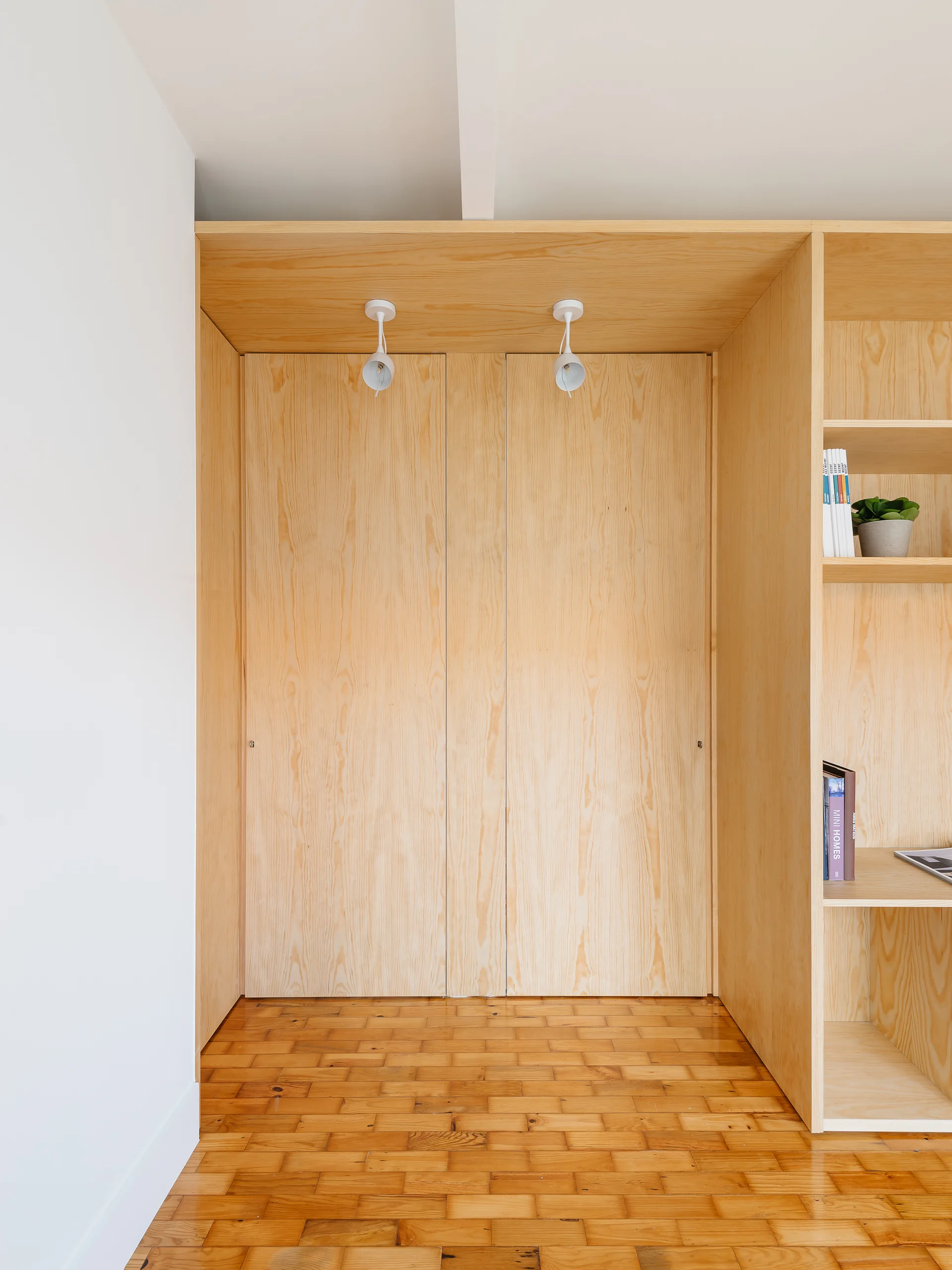 A built-in light wood plywood storage unit with closed doors and open shelves sits above a warm brown parquet wooden floor.