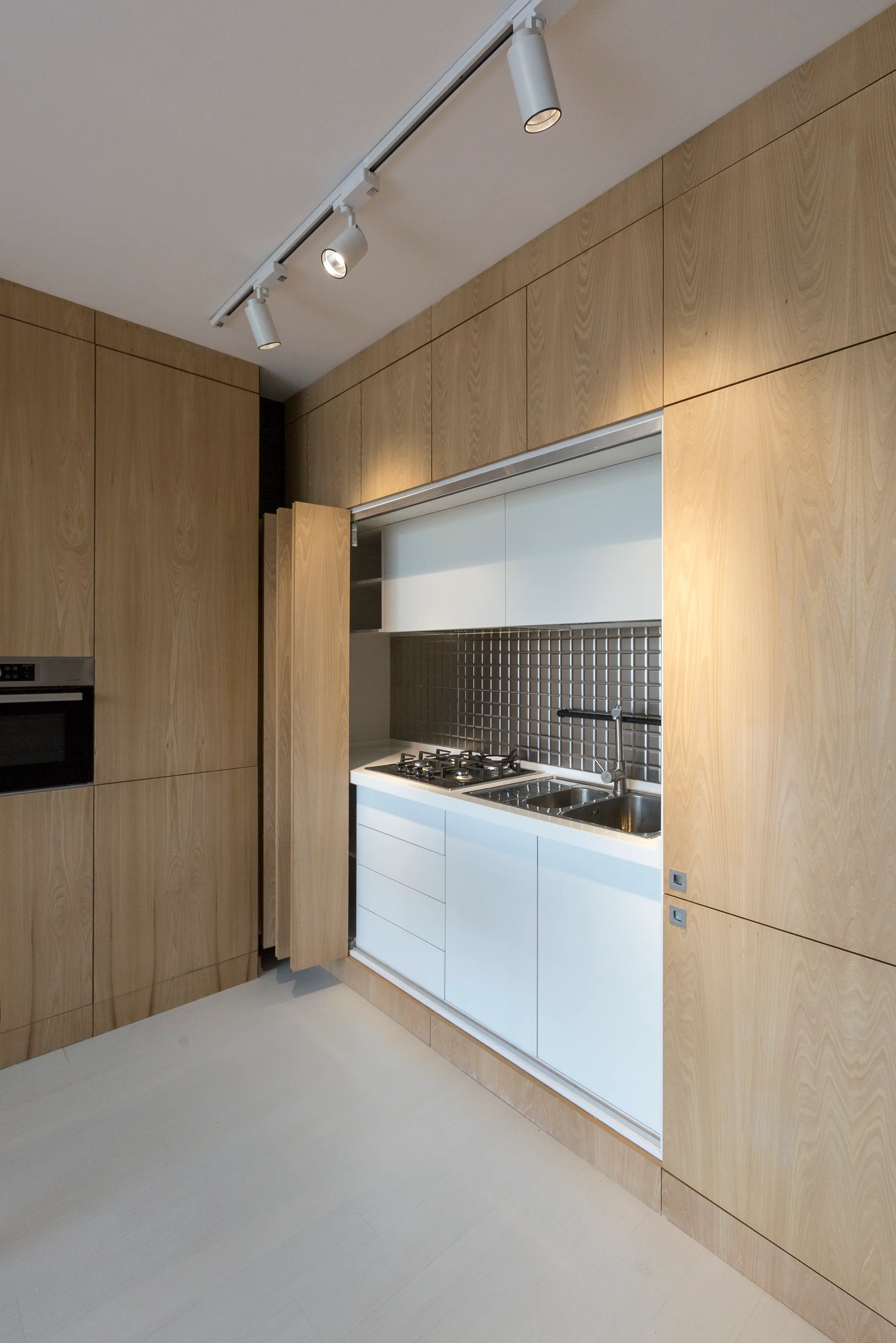 A minimalist concealed kitchenette revealed behind opened light wood bi-fold cabinet doors, featuring white lower cabinets and a metallic tiled backsplash.