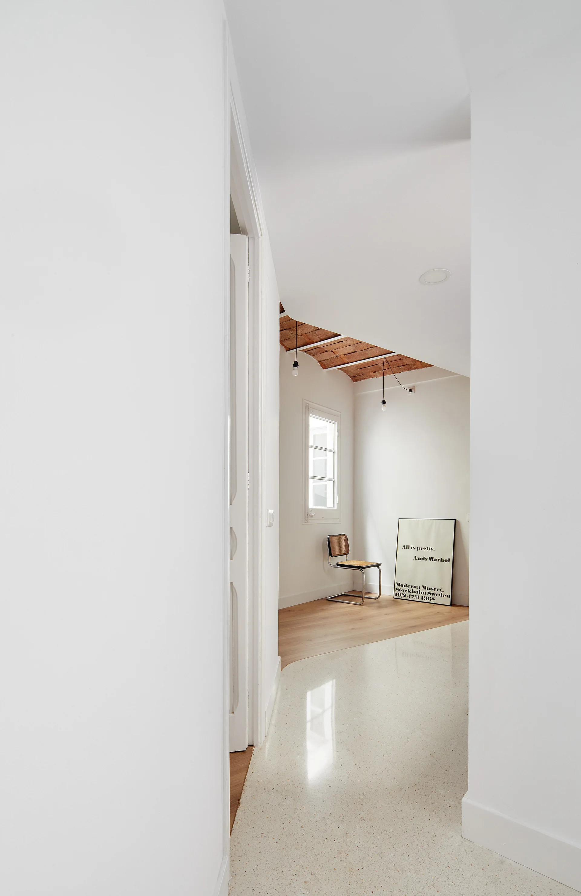 A bright white hallway with polished terrazzo flooring leading into a room with an exposed brick ceiling and a single cantilever chair.